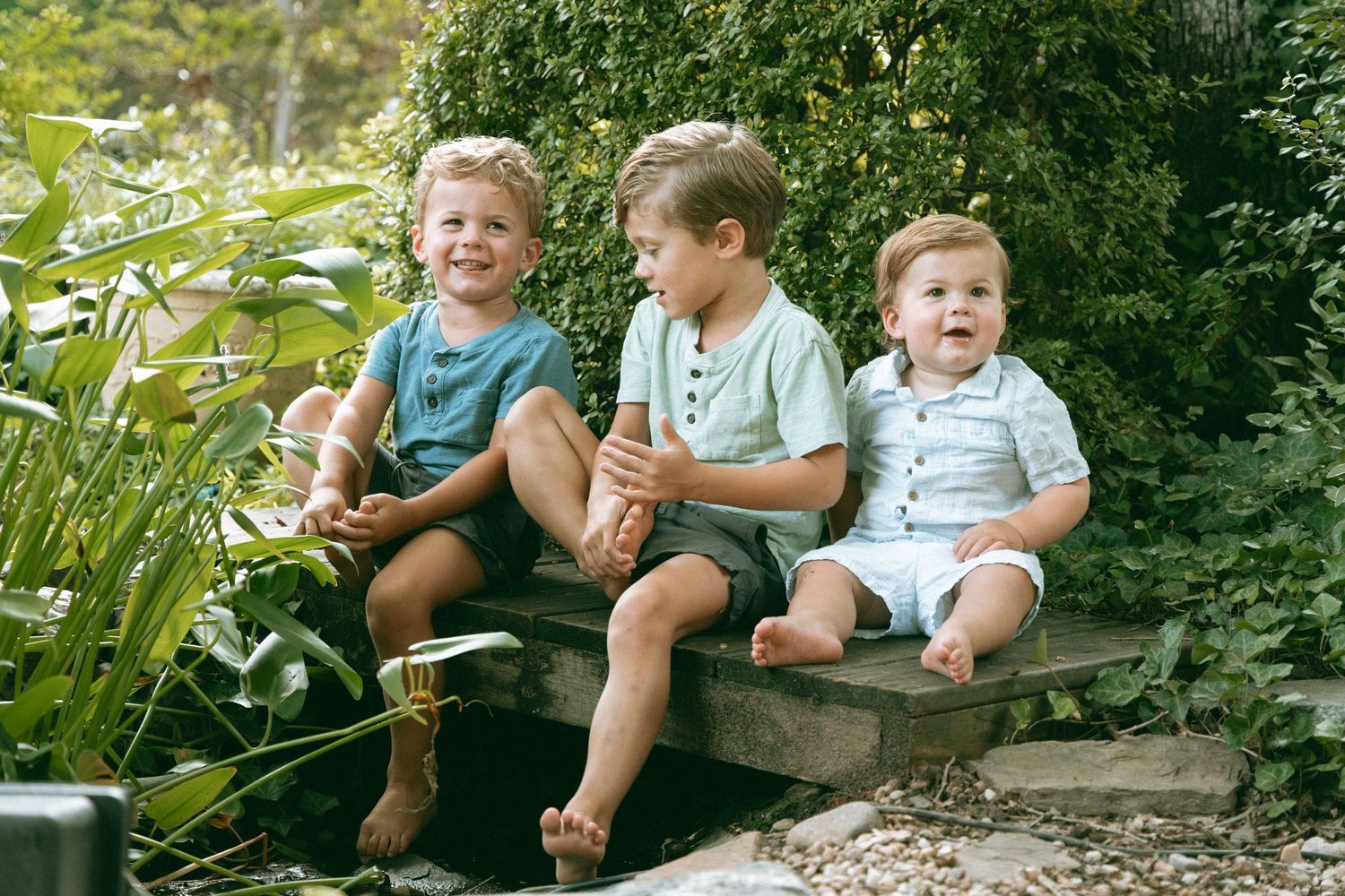 Three young boys sitting together on stone steps surrounded by lush green garden plants, smiling and laughing outdoors.