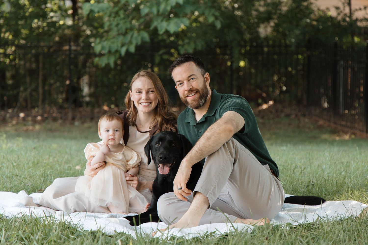 Family of two adults and a baby sitting on a blanket outdoors with a black dog, smiling in a lush green park.