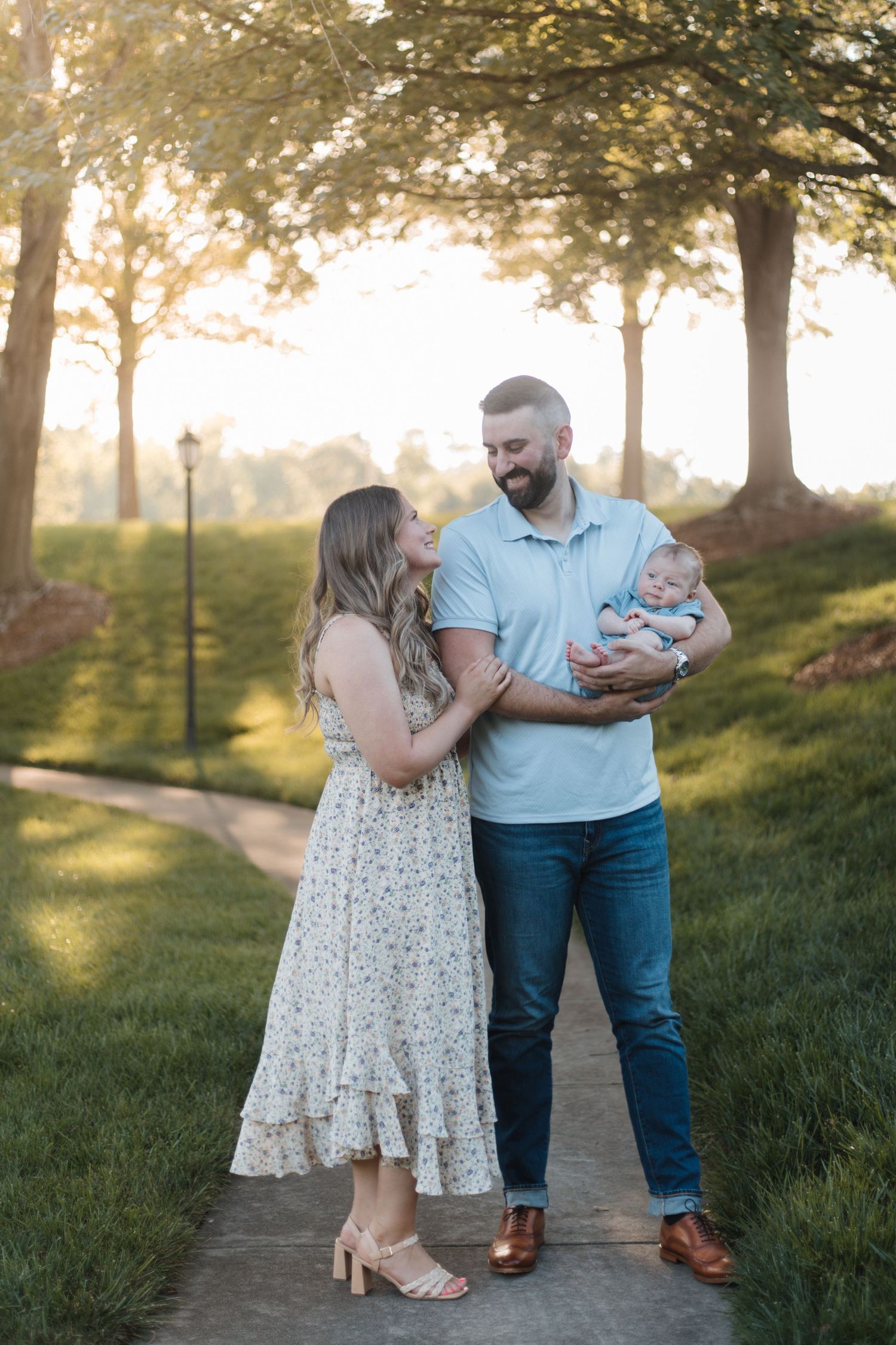 A couple in a sunlit park smiles at their newborn baby, woman in floral dress, man in blue polo and jeans.