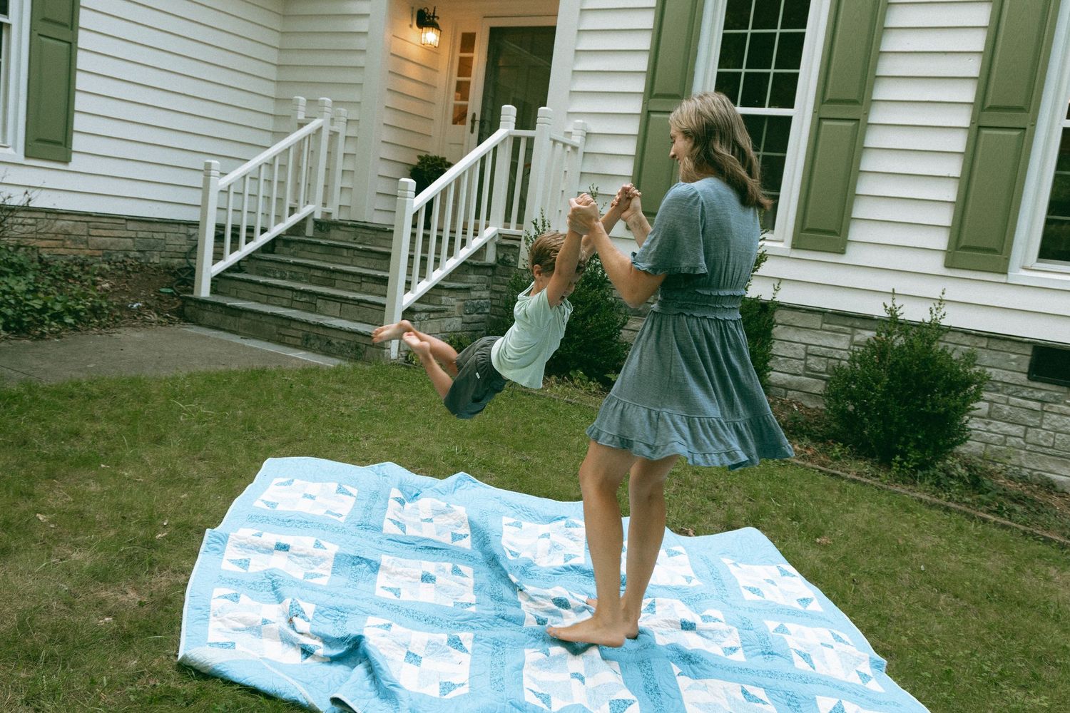 Woman in blue dress swinging toddler over blue patchwork quilt on green lawn outside white house.