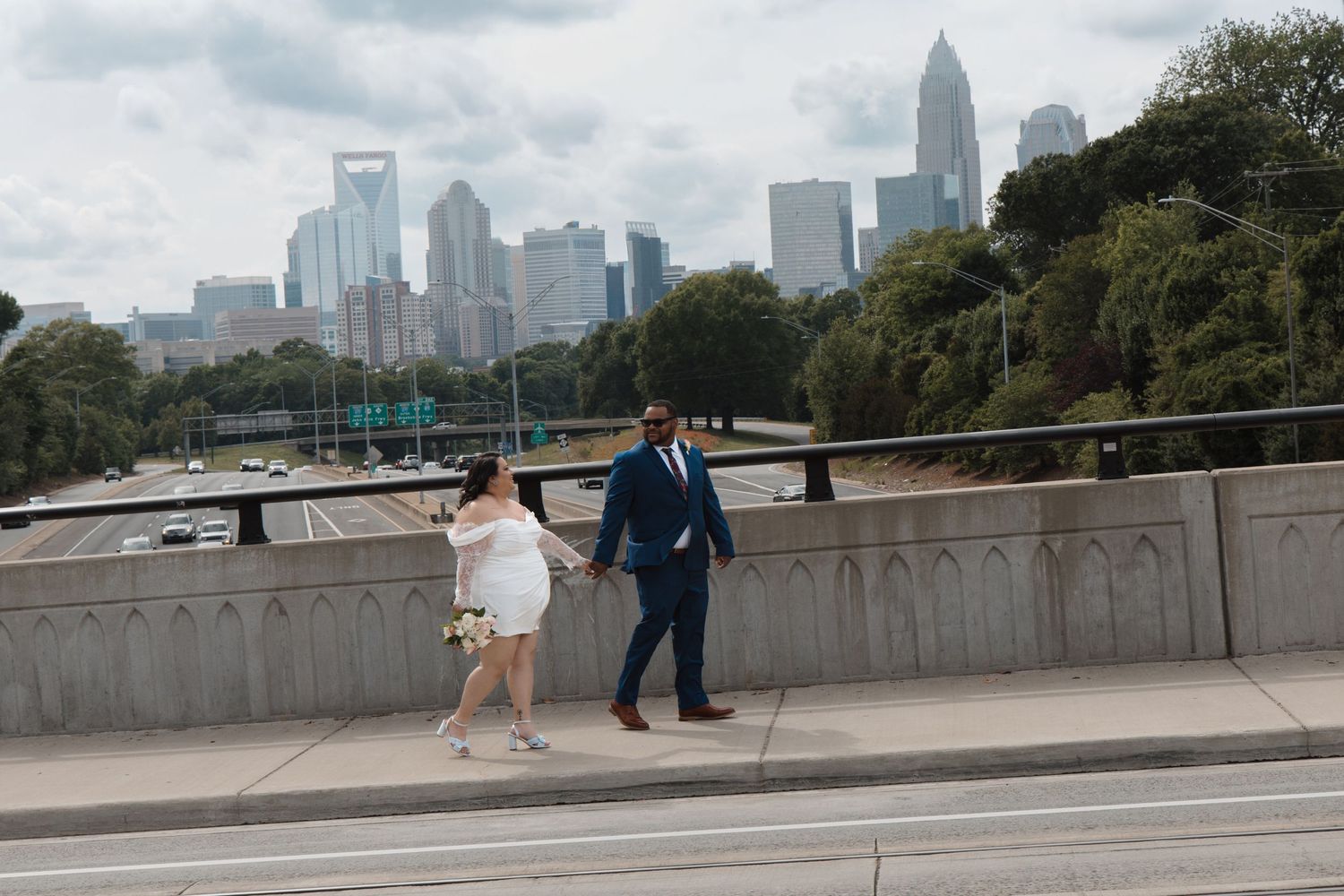 Bride and groom walk hand-in-hand on a bridge with the Charlotte, NC city skyline in the background. CHARLOTTE NC ELOPEMENT PHOTOGRAPHER