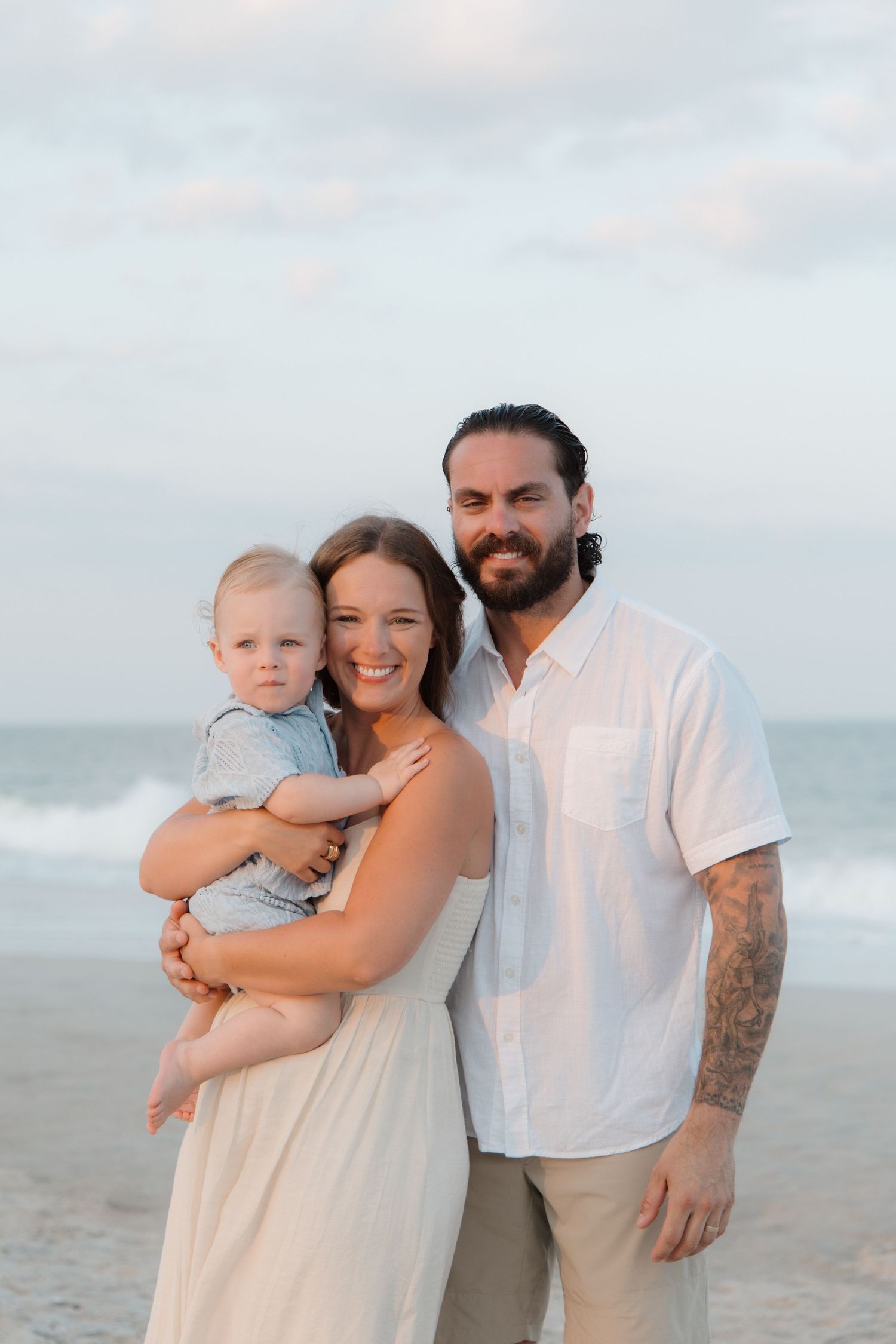 A happy family of three poses on a sandy beach, with a woman in white holding a baby and a tattooed man beside her.