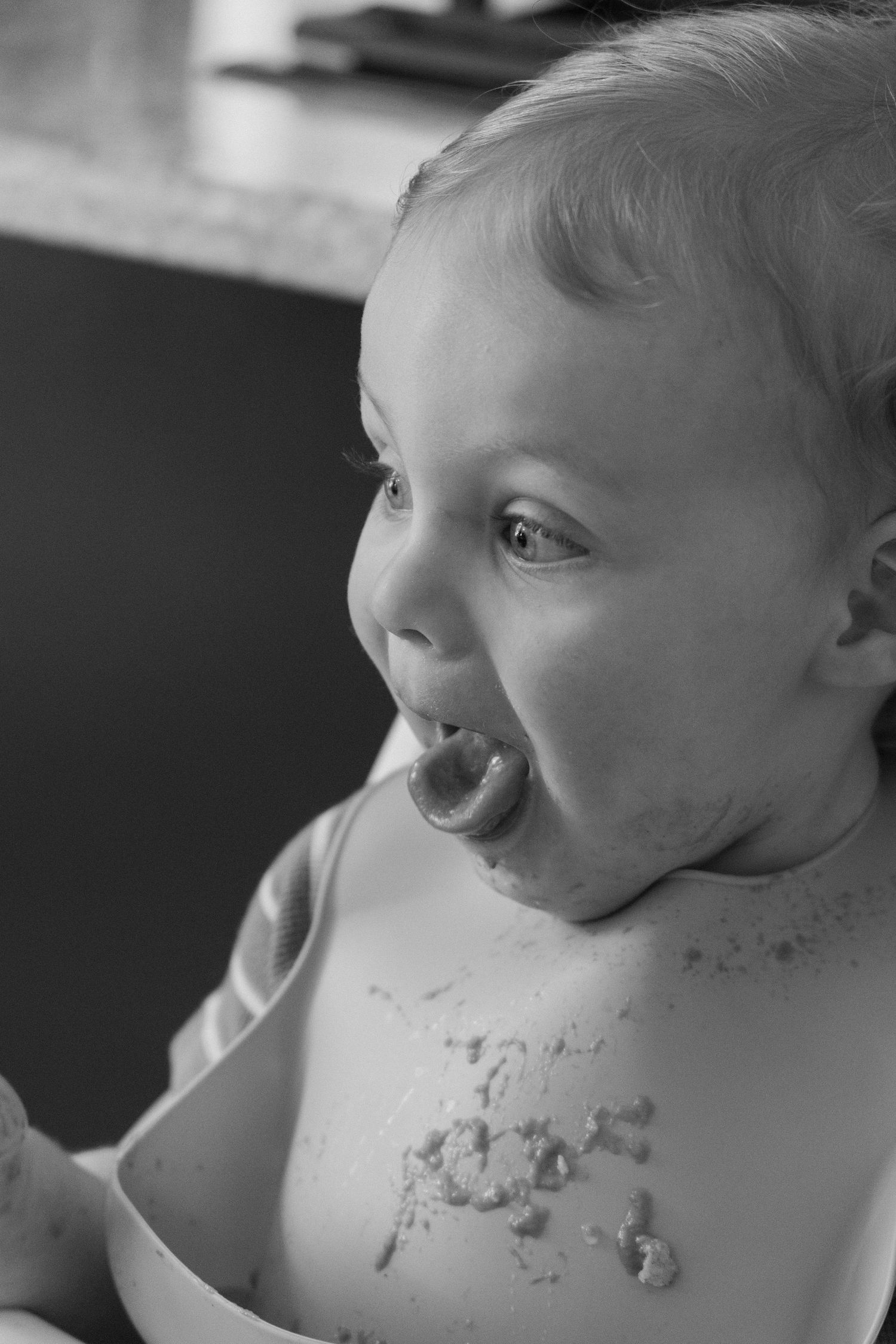 Black and white photo of a happy messy baby with food on chest, mouth open wide, enjoying mealtime.