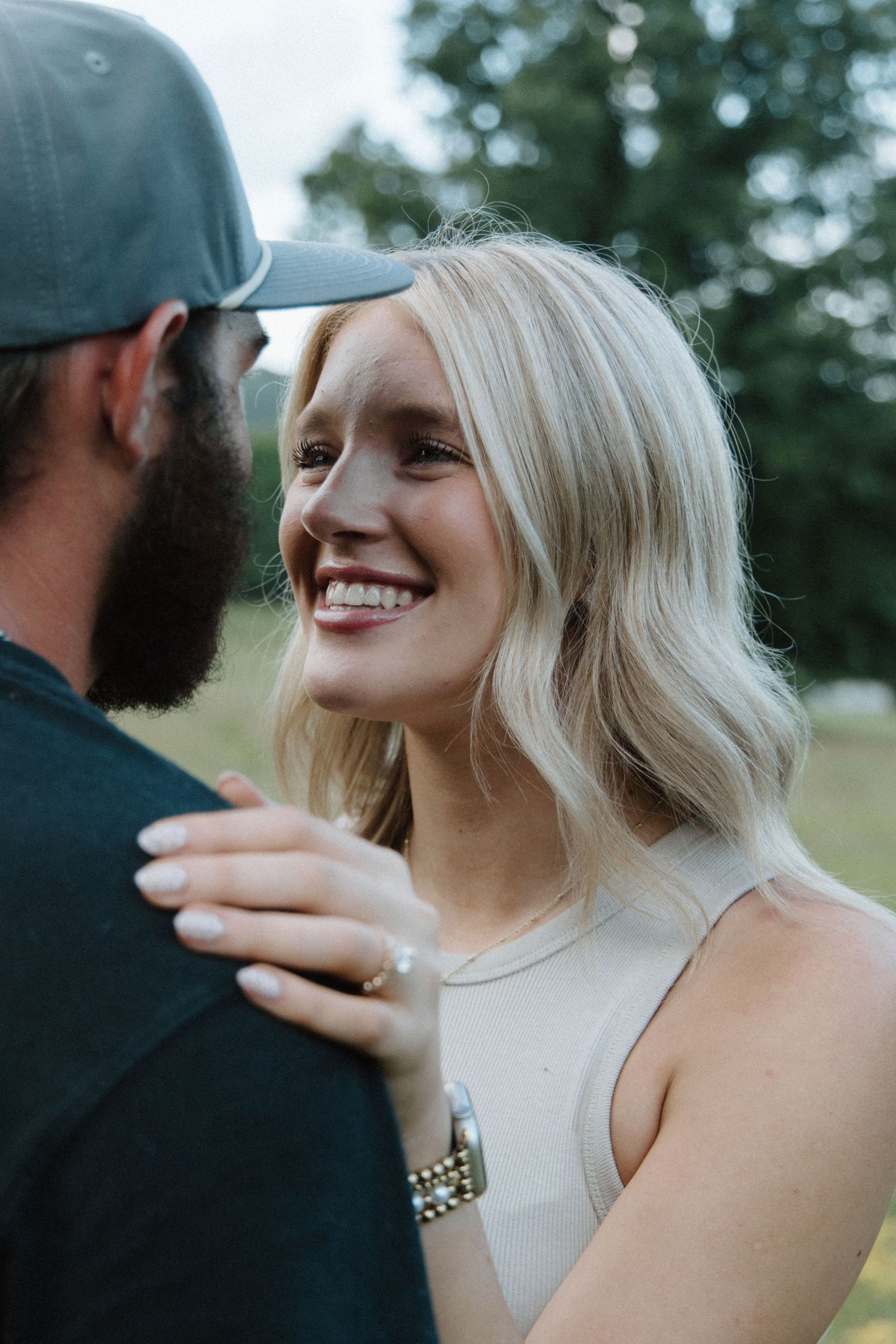 Smiling blonde woman in white top rests her hand on a bearded man's shoulder outdoors near green trees.