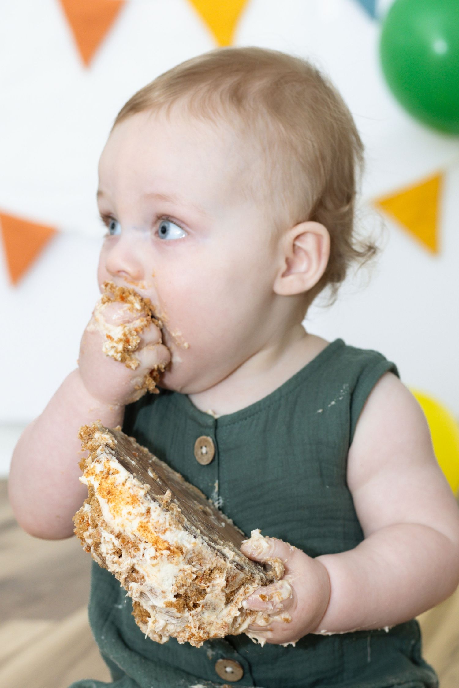 Baby in green romper joyfully smashing and eating first birthday cake with colorful party decorations in background.