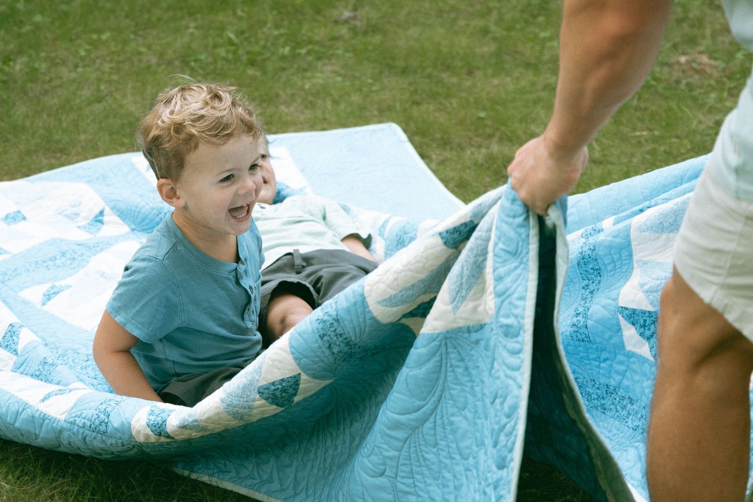 Young boy laughing on a light blue quilt outdoors while an adult playfully wraps him in the blanket.