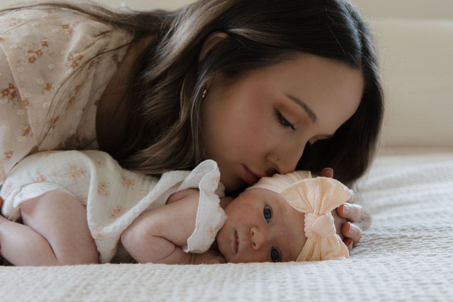 A mother tenderly kisses her newborn baby lying on a soft white blanket wearing a bow headband.
