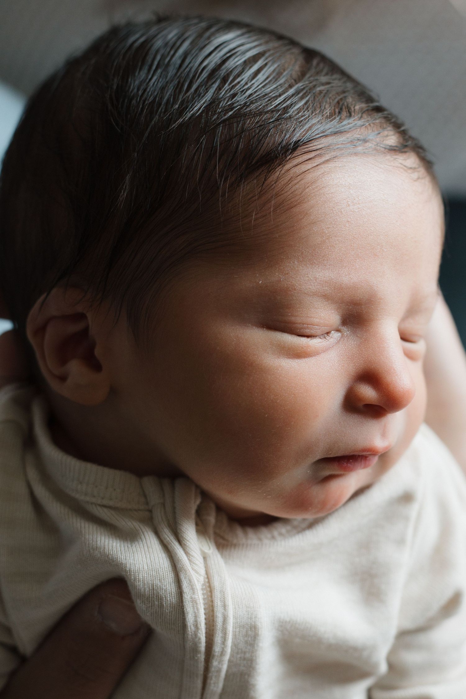Close-up portrait of a peaceful sleeping newborn baby with dark hair wearing a cream onesie. CHARLOTTE NC