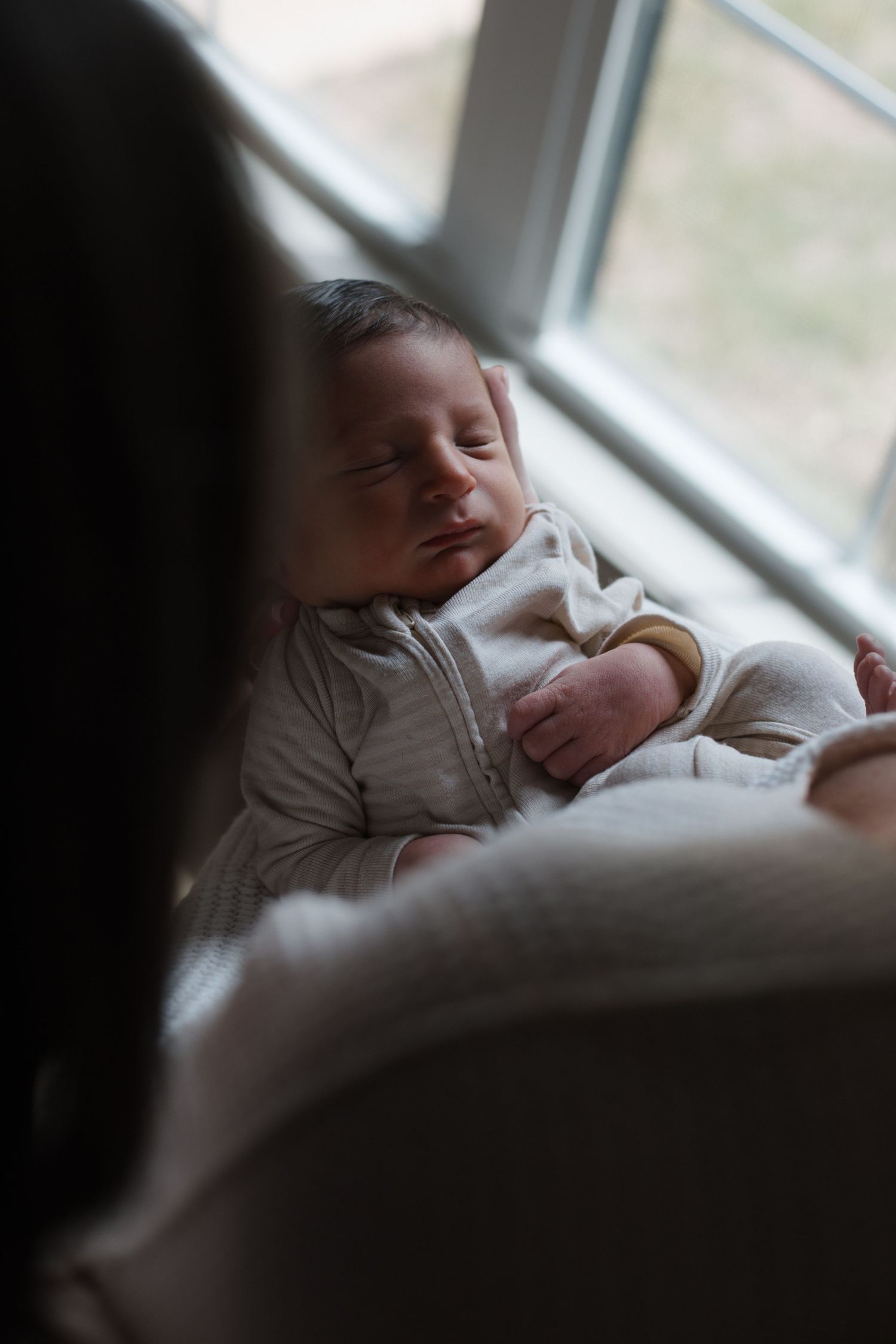 A peaceful newborn baby sleeps in white onesie while being held near a bright window.