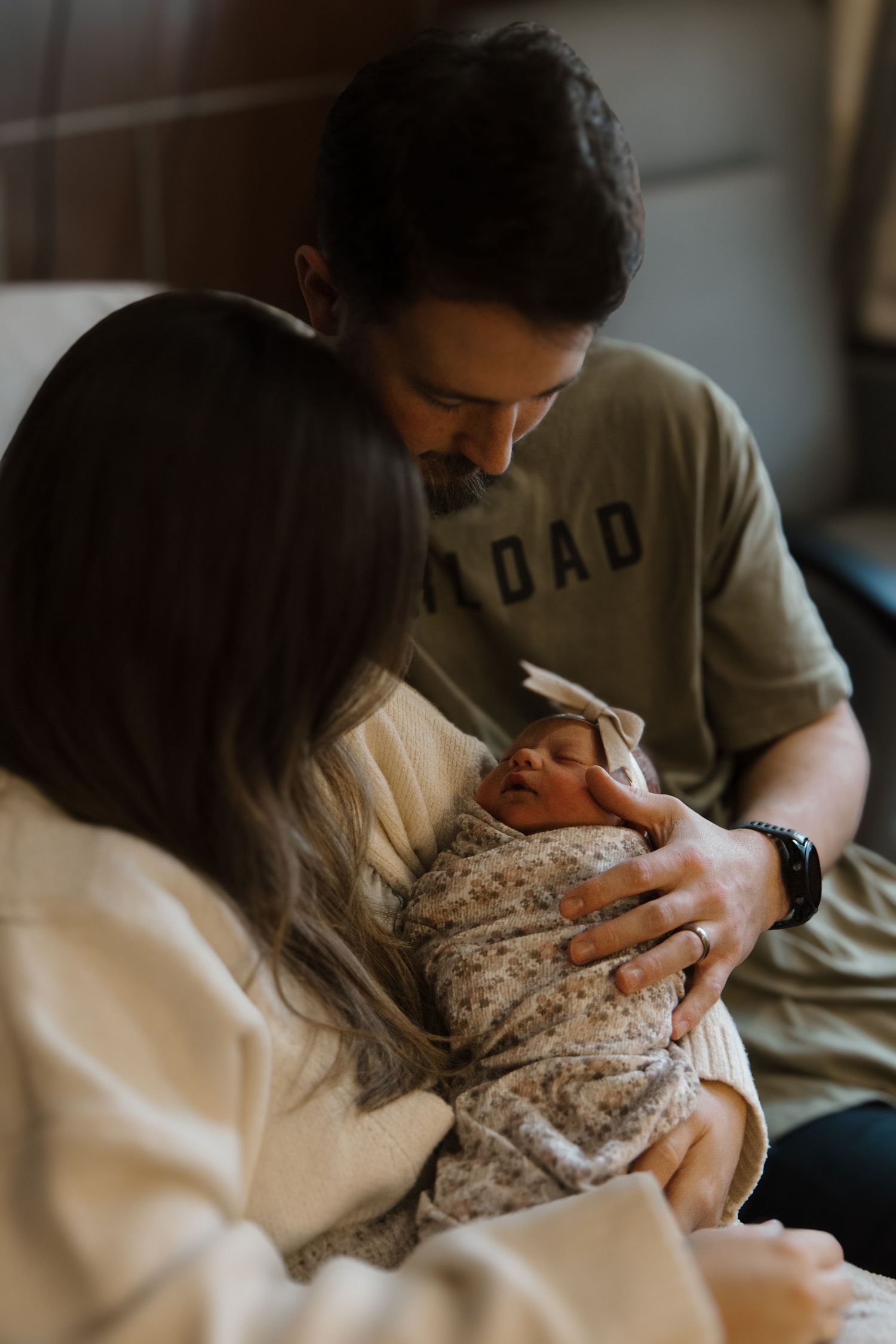 New parents tenderly holding their swaddled newborn baby, father wearing a DAD shirt, in a warm hospital room. Charlotte NC Matthews NC
