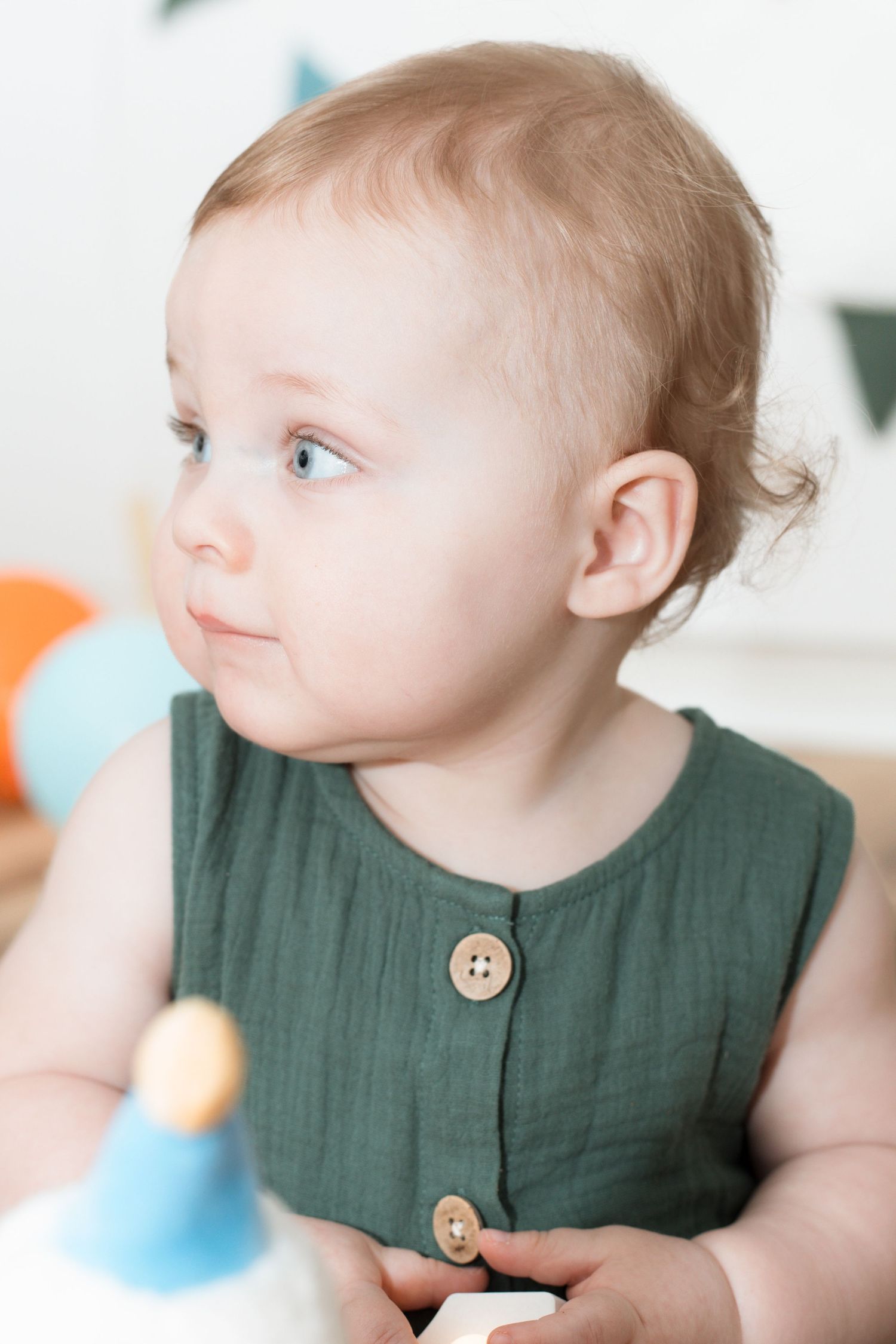 Adorable baby with blue eyes wearing a green linen romper, looking sideways during a birthday celebration.