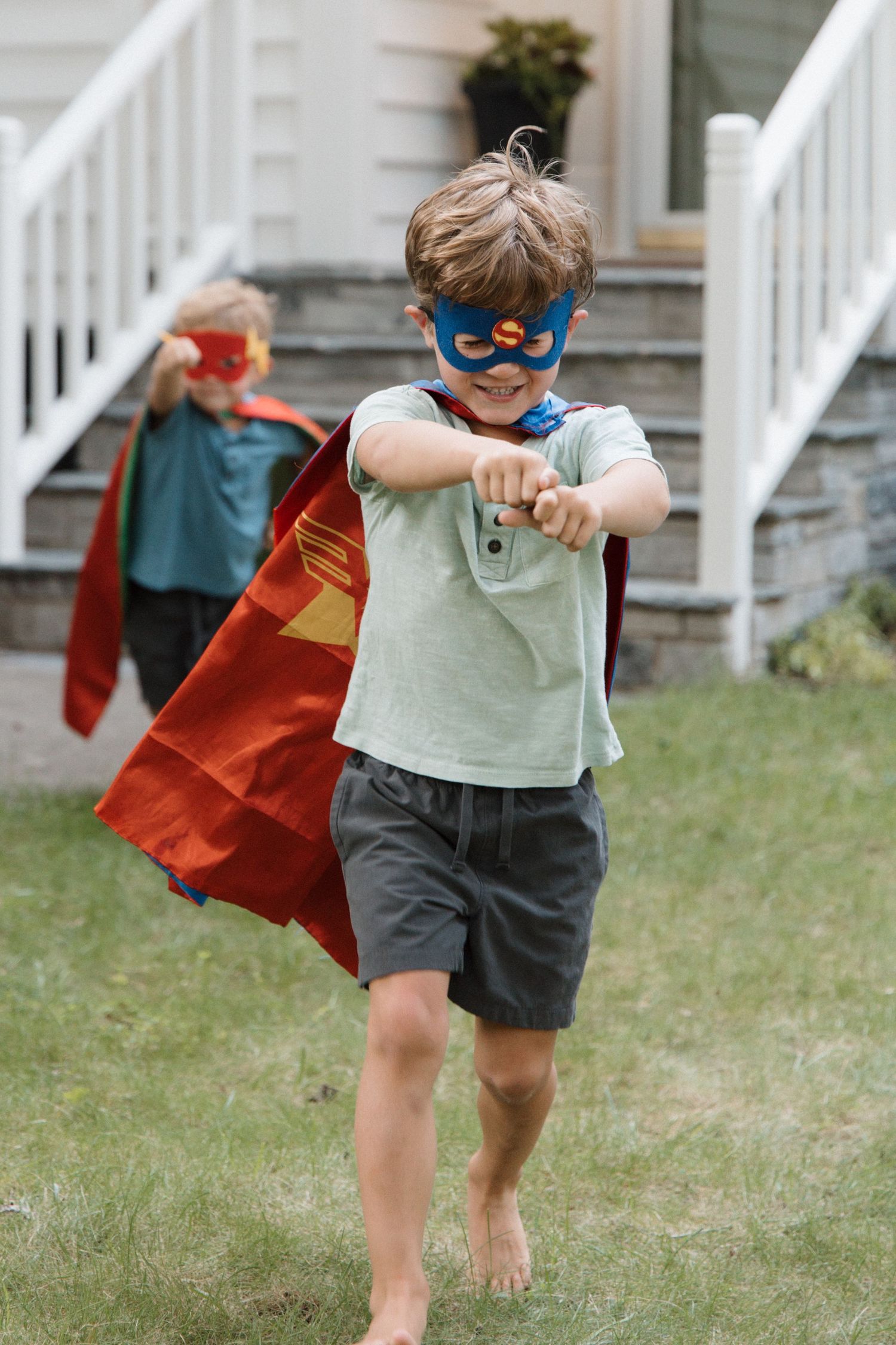 Two young boys dressed in superhero capes and masks play pretend outside a house on a sunny day.