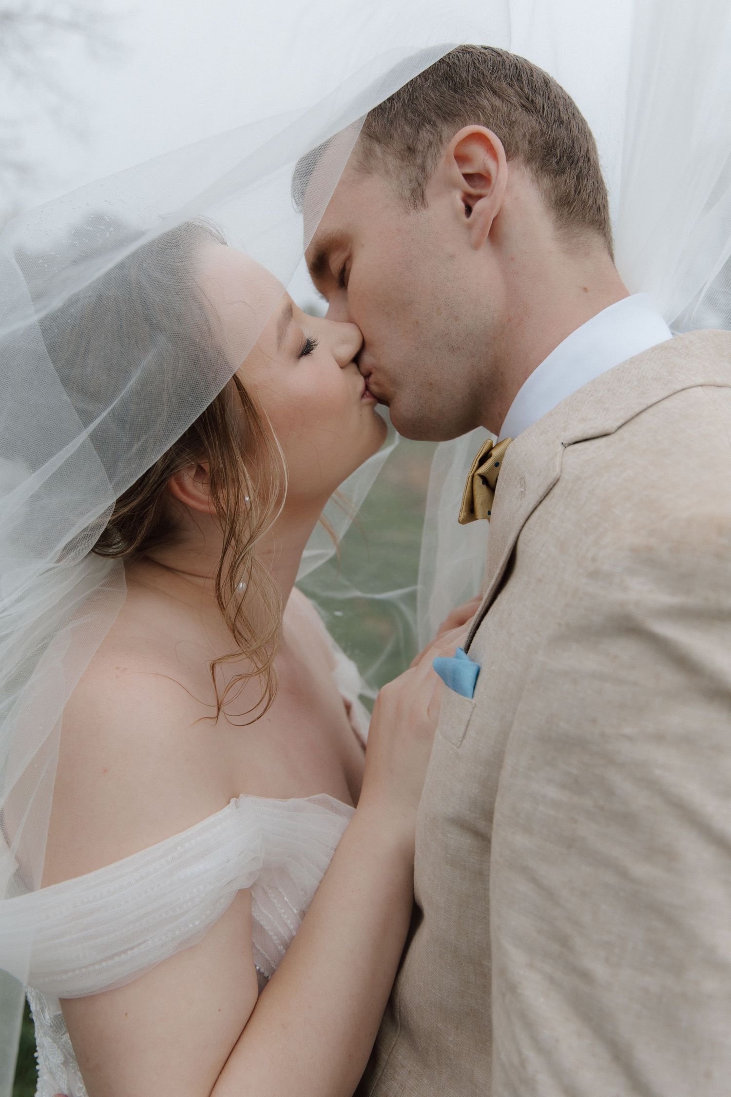 Bride and groom sharing a romantic kiss under a flowing wedding veil, groom in beige suit with blue boutonniere. CHARLOTTE NC