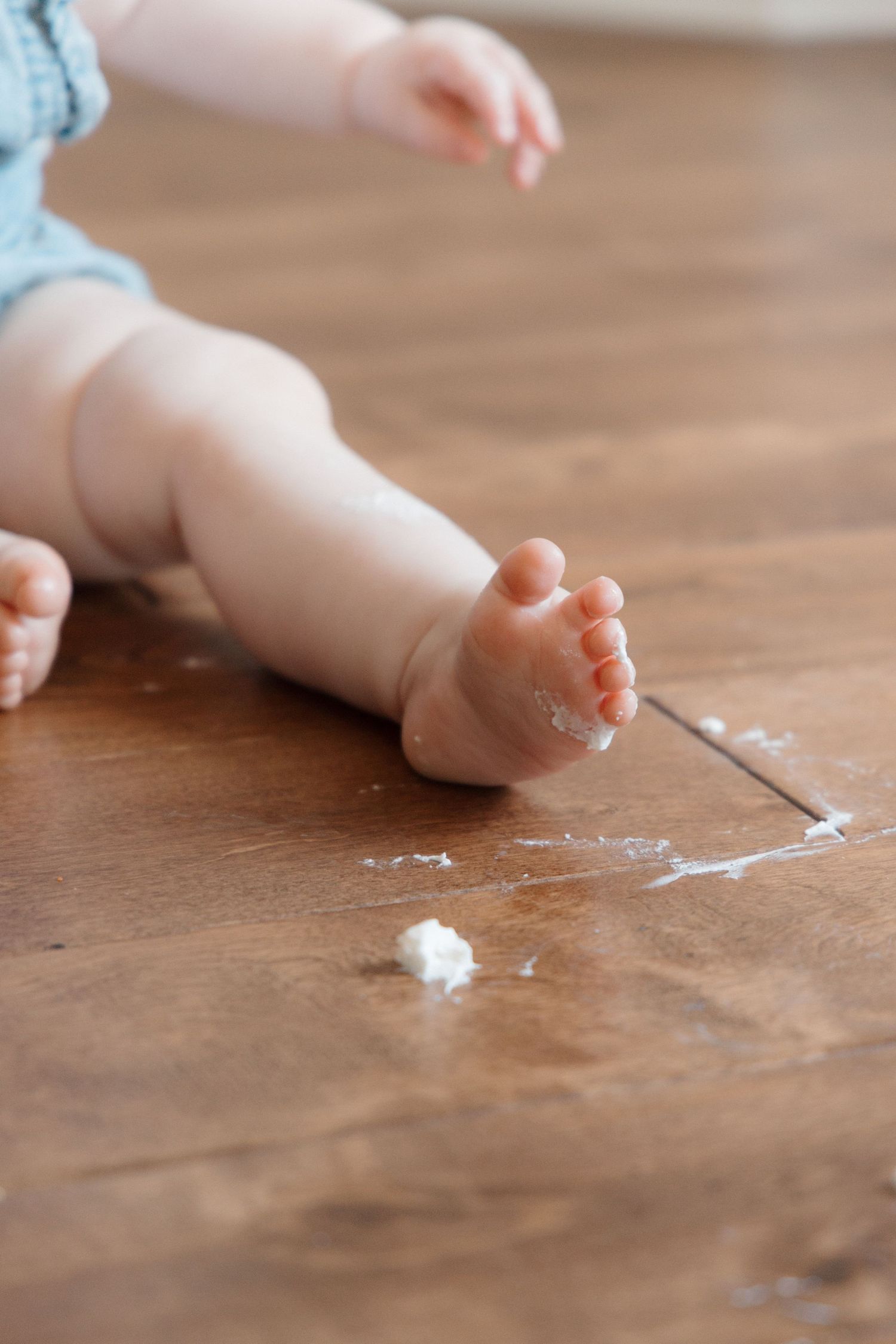 Close-up of chubby baby feet sitting on wooden floor with smeared white frosting during first birthday cake smash.