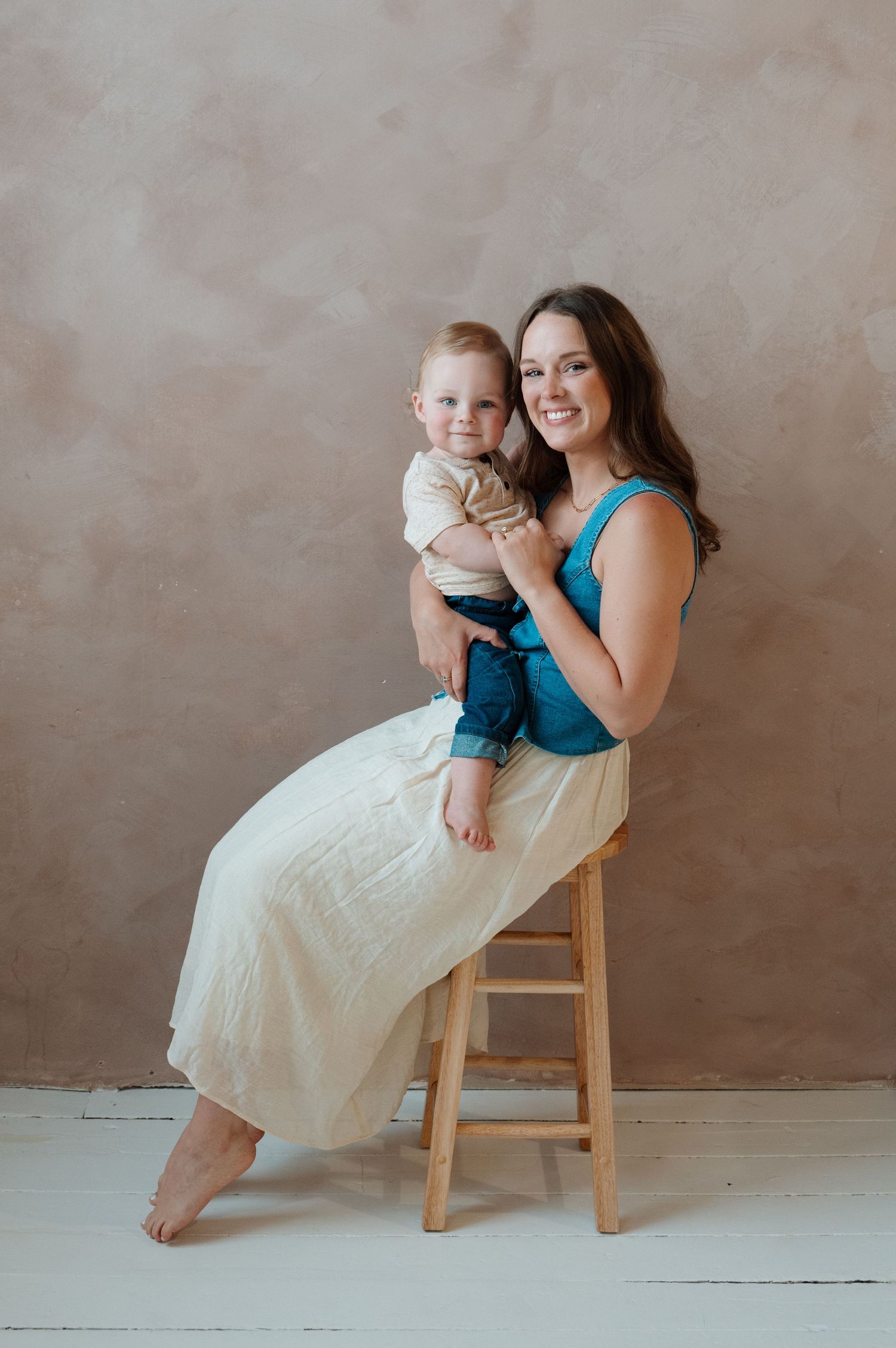 A smiling woman in a teal top and cream skirt sits on a wooden stool holding a baby in a photo studio.