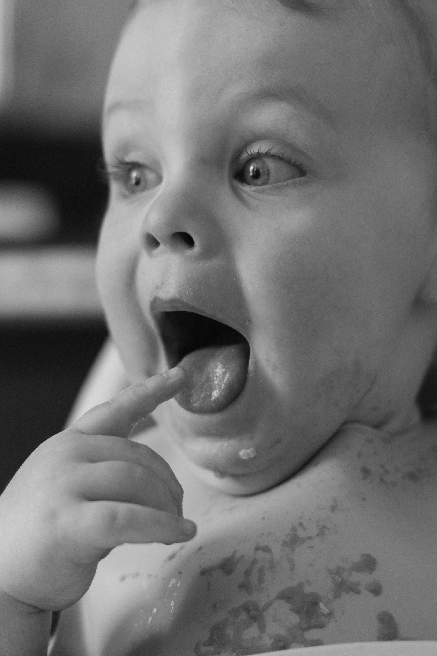 Black and white photo of a messy baby licking their finger with wide eyes and an open mouth.