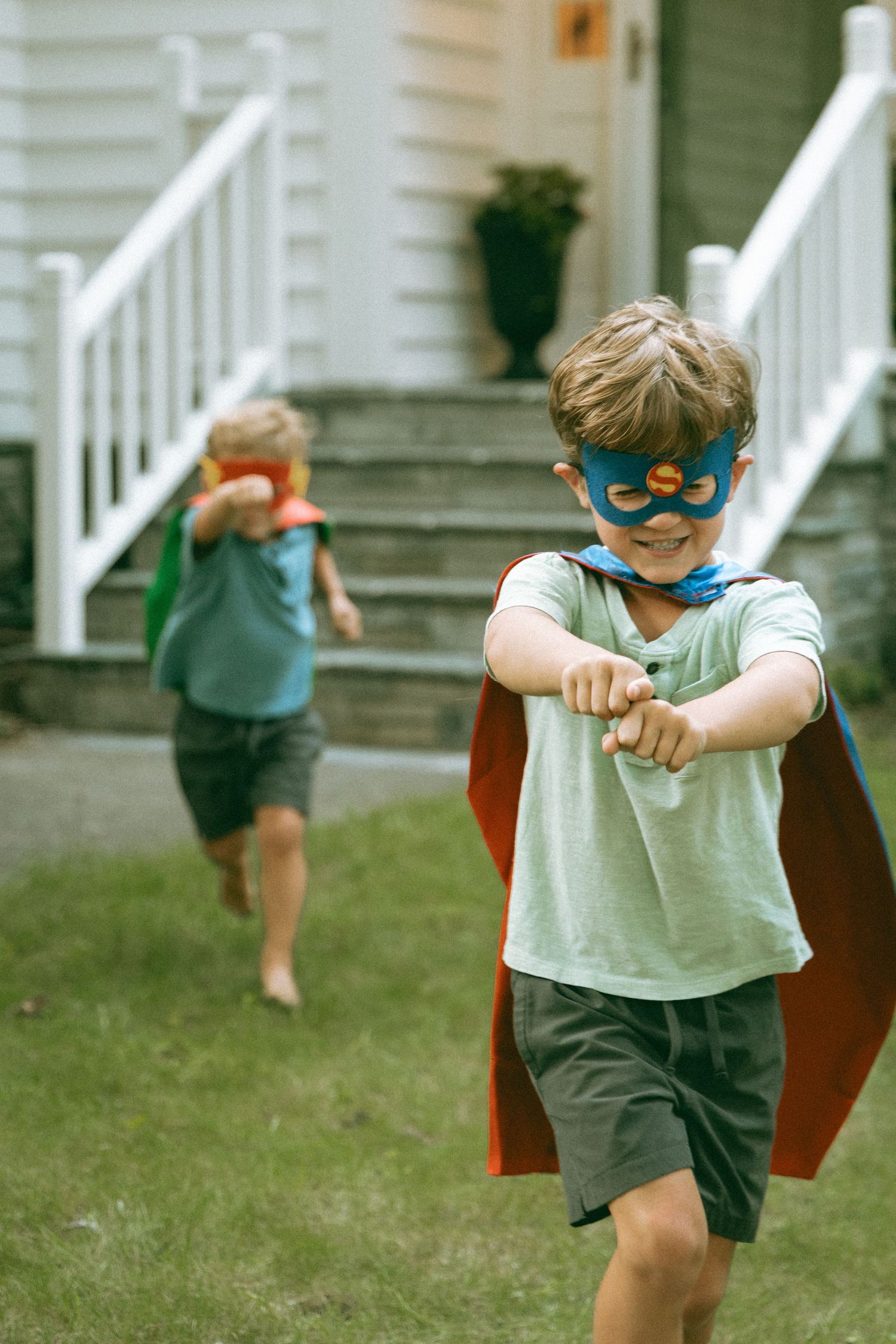 Two boys in superhero capes and masks play pretend outside a house on a sunny day.