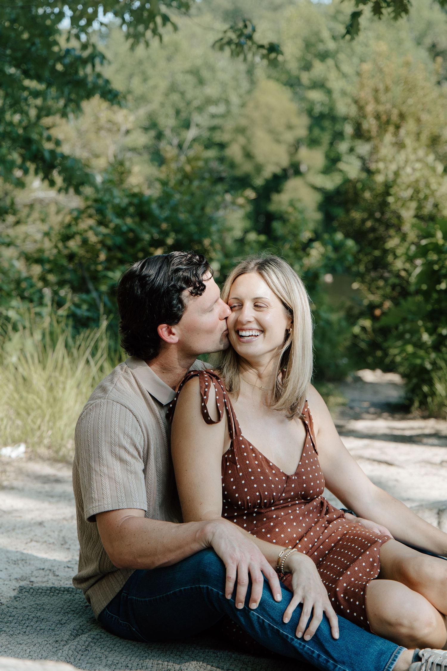 A couple sits outdoors, man kissing woman's cheek as she smiles, surrounded by lush green trees.