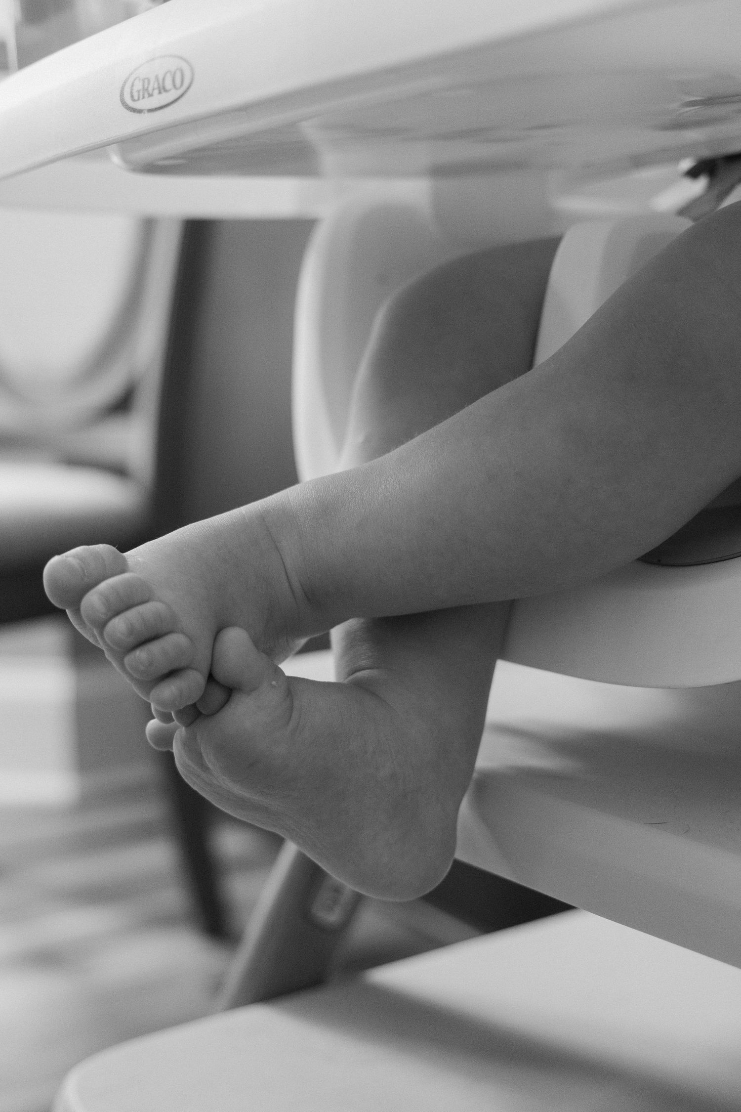 Black and white close-up of tiny baby feet dangling from a Graco high chair.