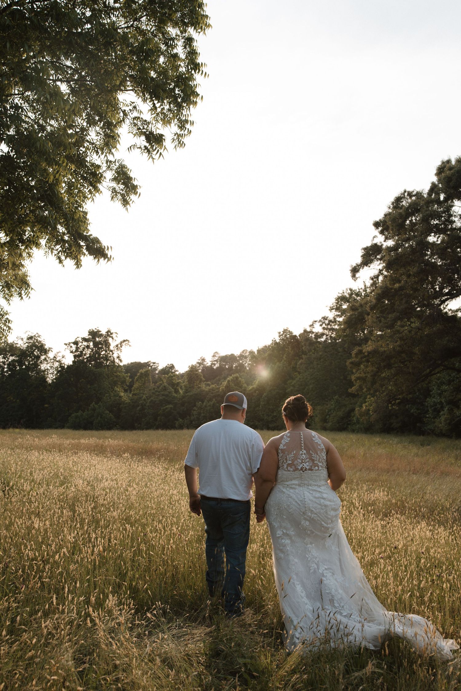 Bride and groom walking hand in hand through a golden field at sunset, surrounded by lush green trees.