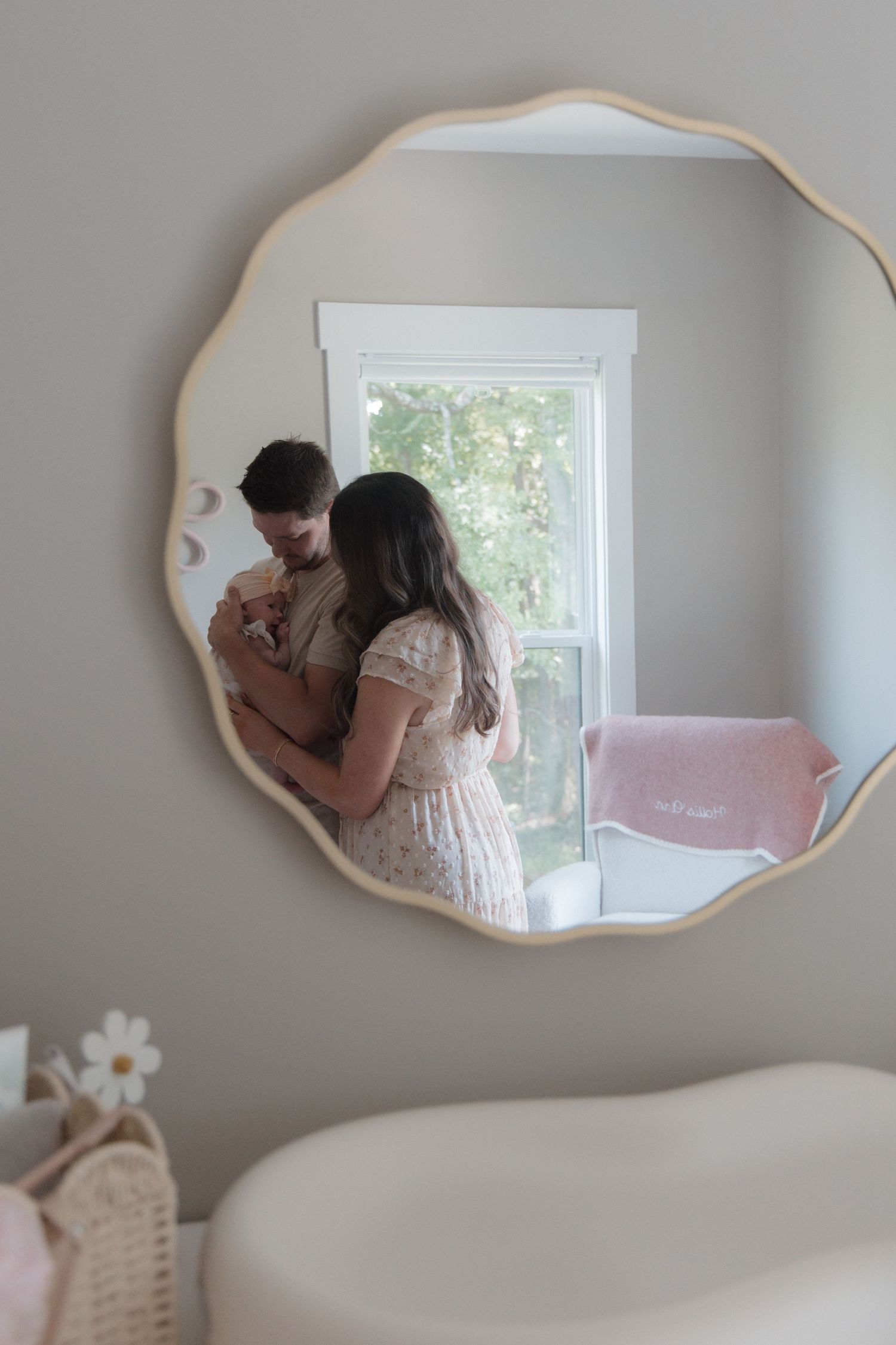 Mother holding newborn baby reflected in decorative wavy gold mirror in soft neutral nursery room.
