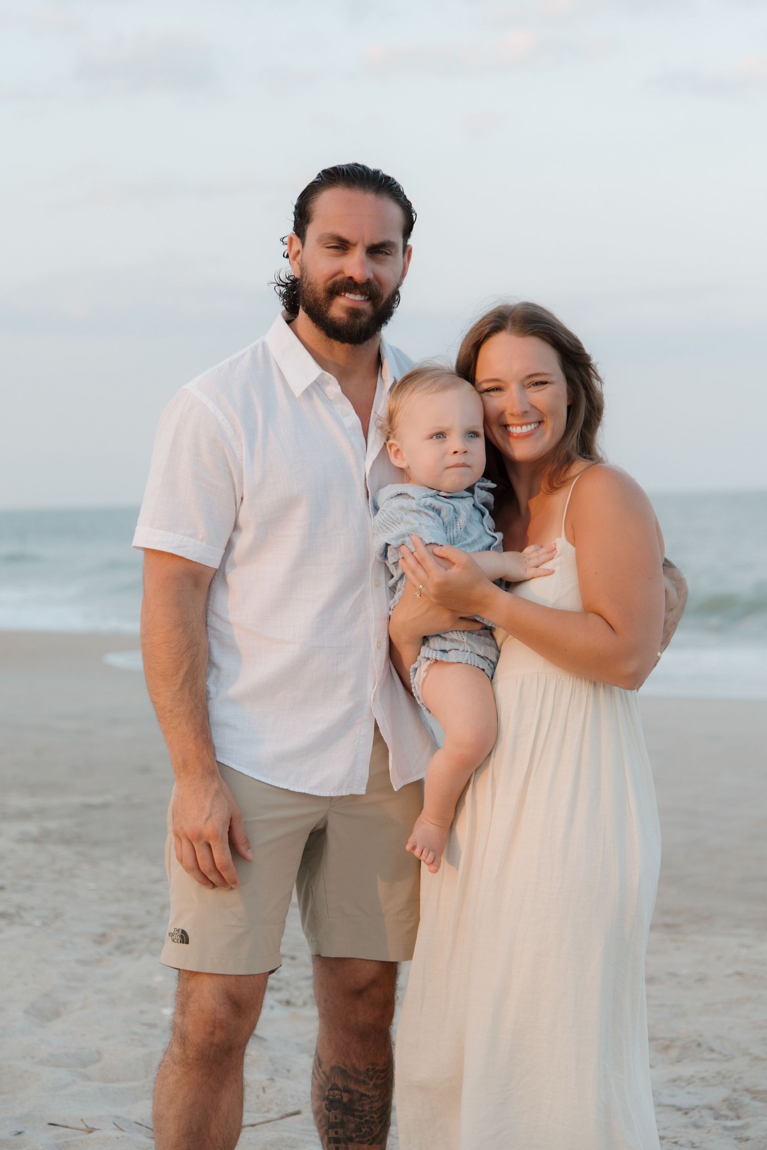 A smiling couple stands on a beach holding their baby, dressed in white and khaki, with ocean waves in the background.