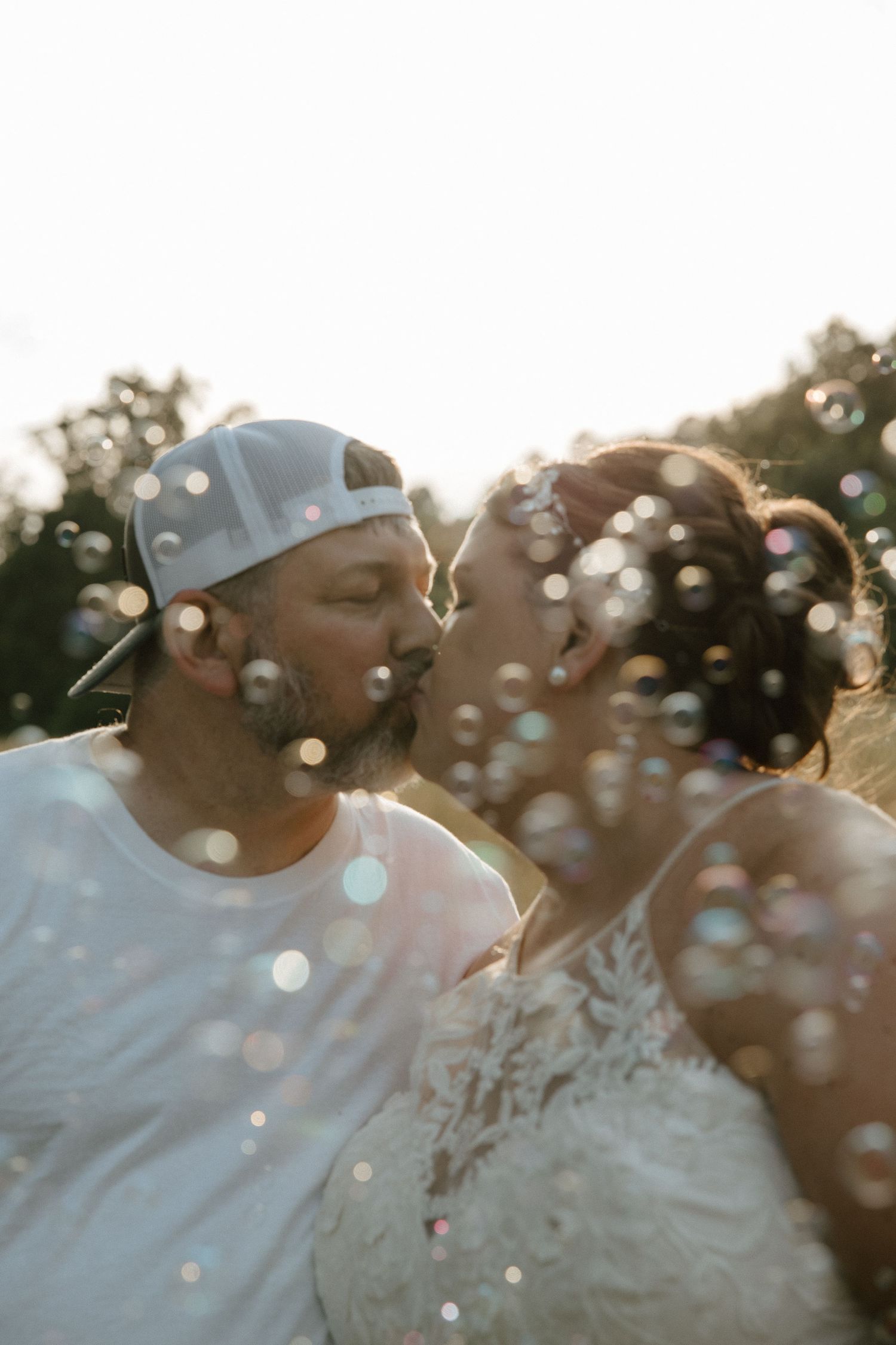 A couple shares a romantic kiss surrounded by floating bubbles during a golden hour outdoor wedding celebration.