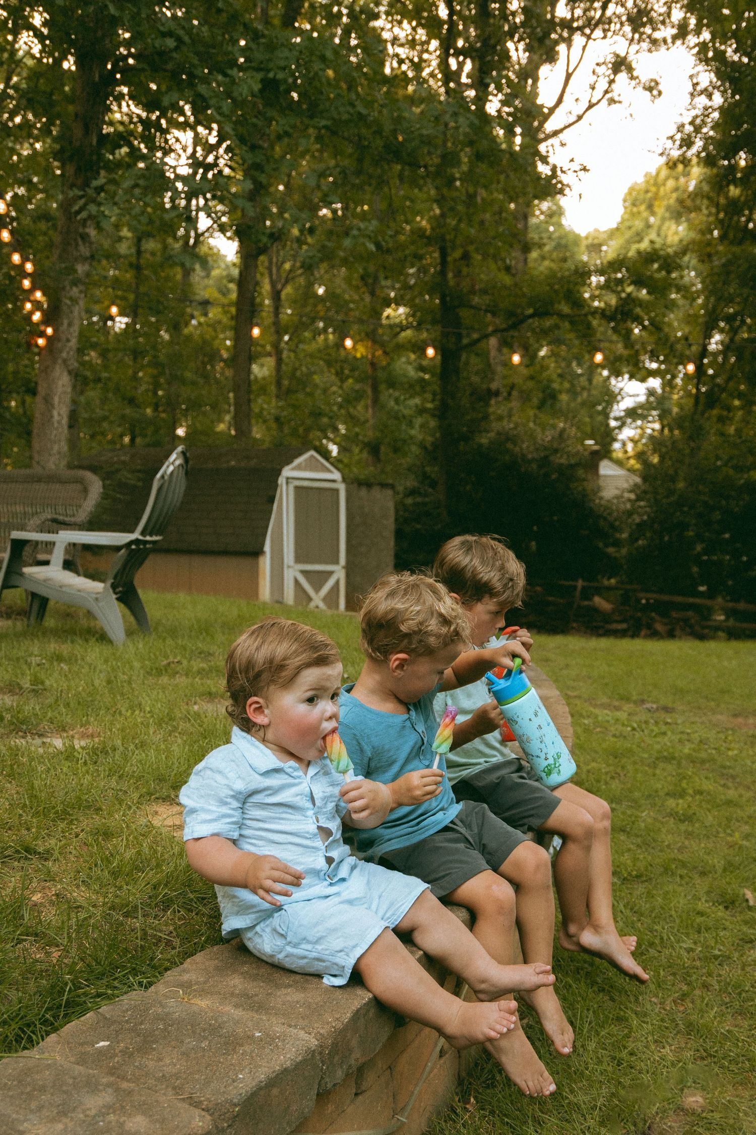 Three young boys sitting on a stone ledge in a backyard, blowing bubbles and enjoying popsicles at dusk.