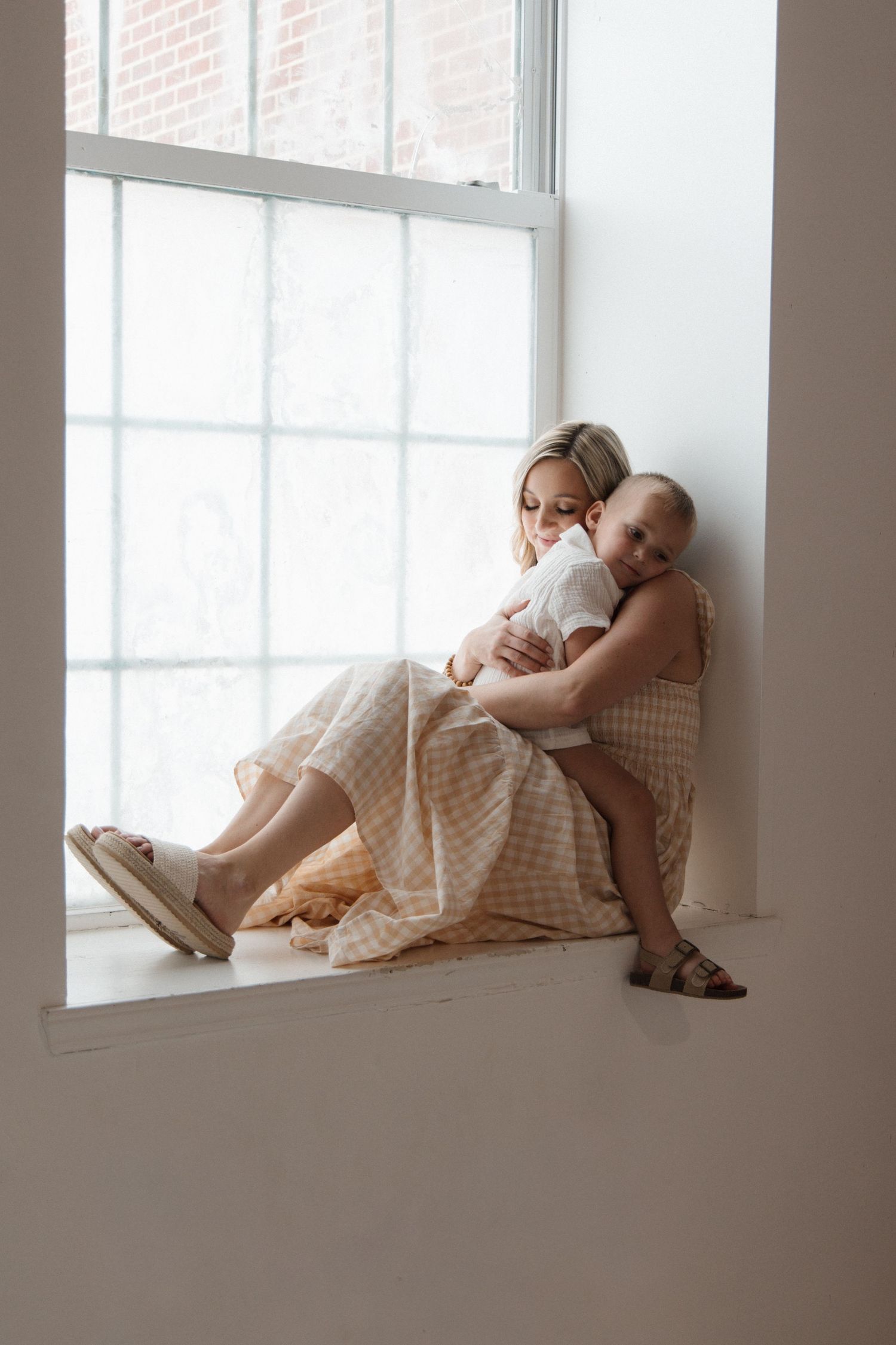 A woman in a beige gingham dress sits on a windowsill, tenderly holding a baby in soft natural light.
CHARLOTTE NC PHOTOGRAPHER
