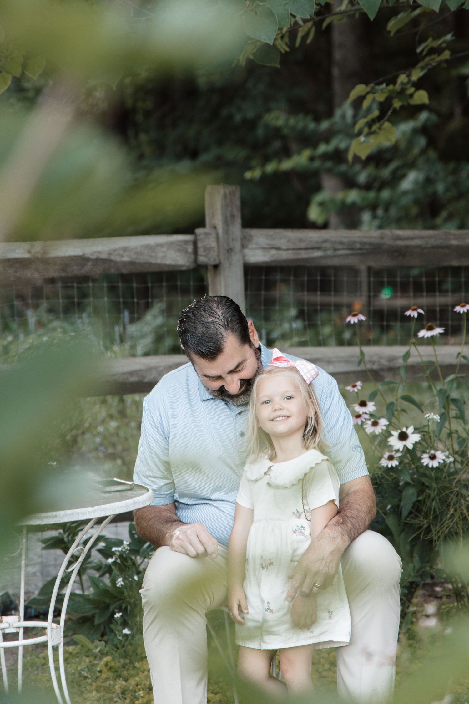 Father and daughter share a tender moment in a lush garden, sitting together near wooden fence with blooming flowers.