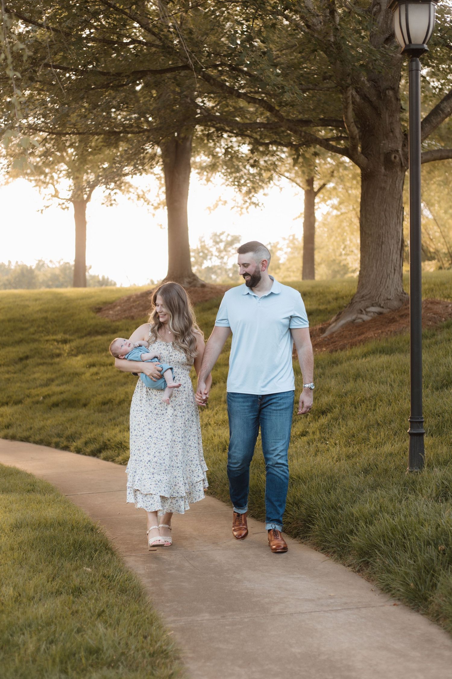 A couple walks along a park path at golden hour, with the woman holding a newborn baby.