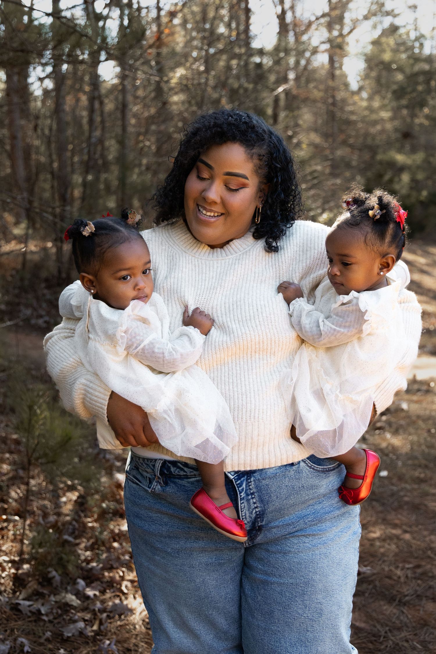 A smiling mother holds her twin toddler daughters in a sunlit forest, all dressed in white outfits.