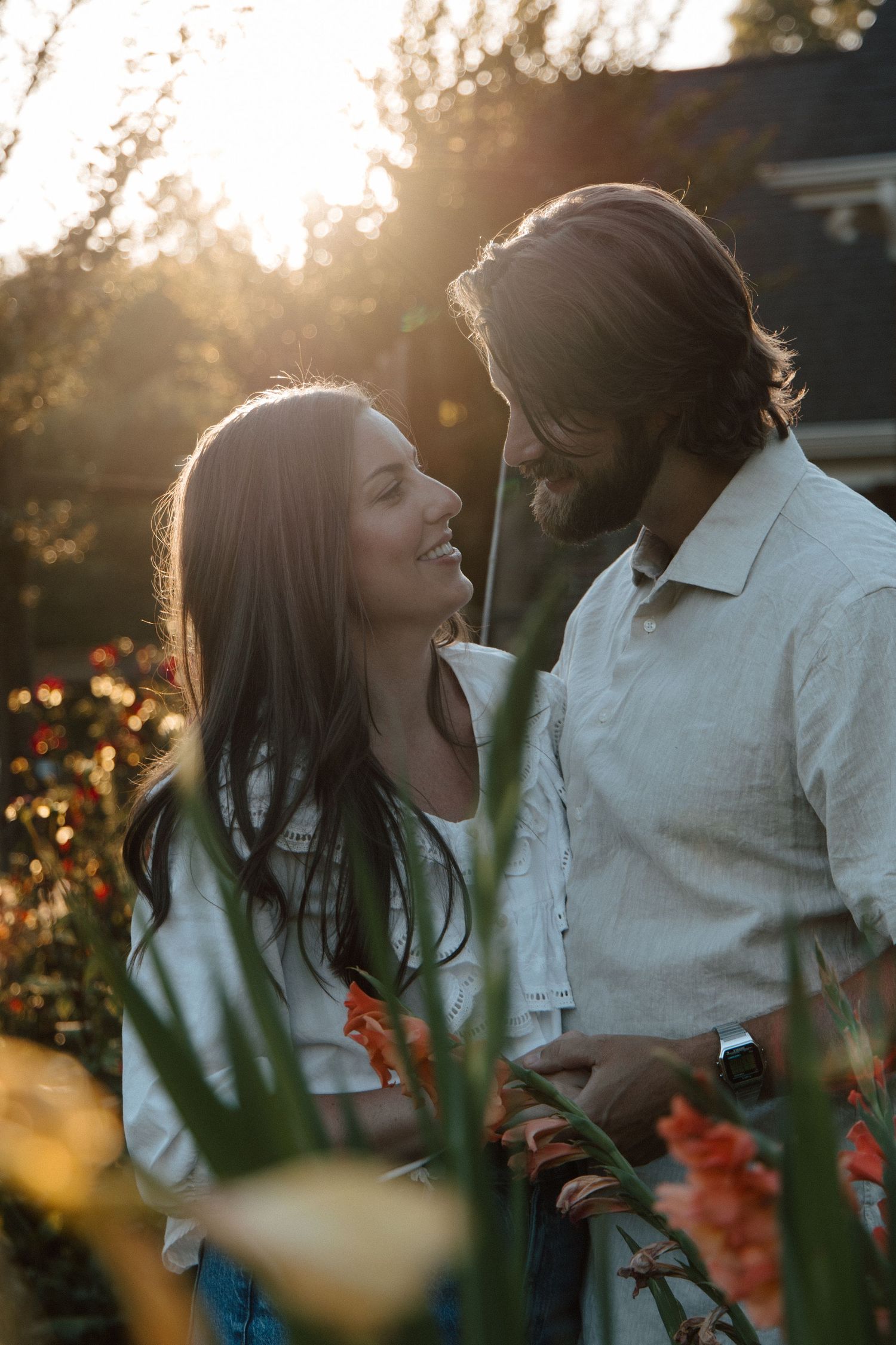 Couple smiling at each other in a sunlit garden with colorful flowers during golden hour.