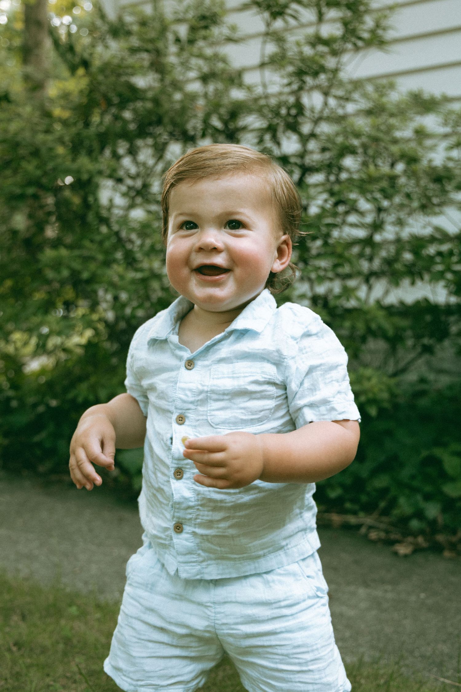 Smiling baby boy in white linen outfit standing outdoors in a green garden.