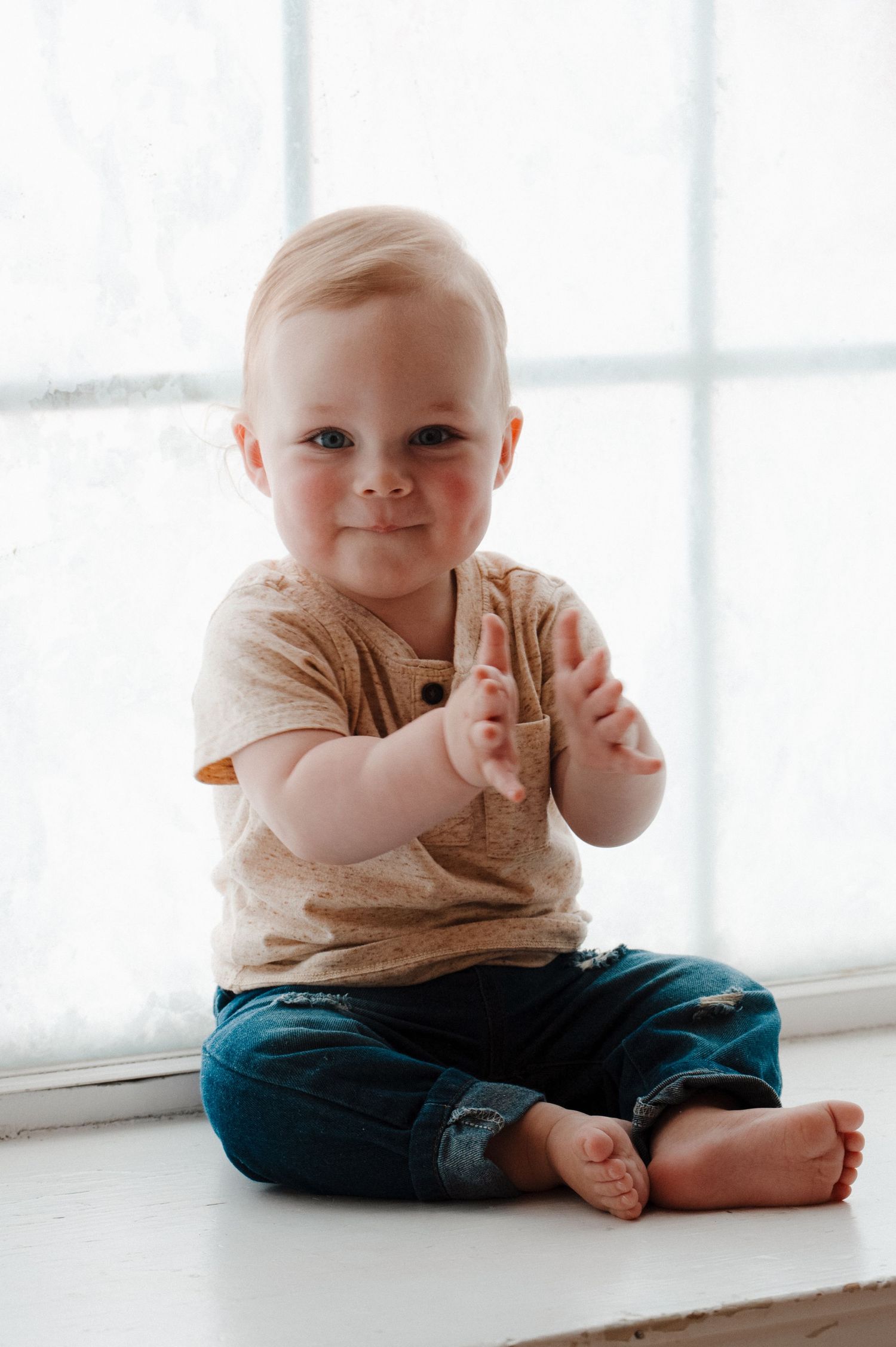 Smiling baby in beige shirt and jeans sitting cross-legged on white windowsill, clapping hands.