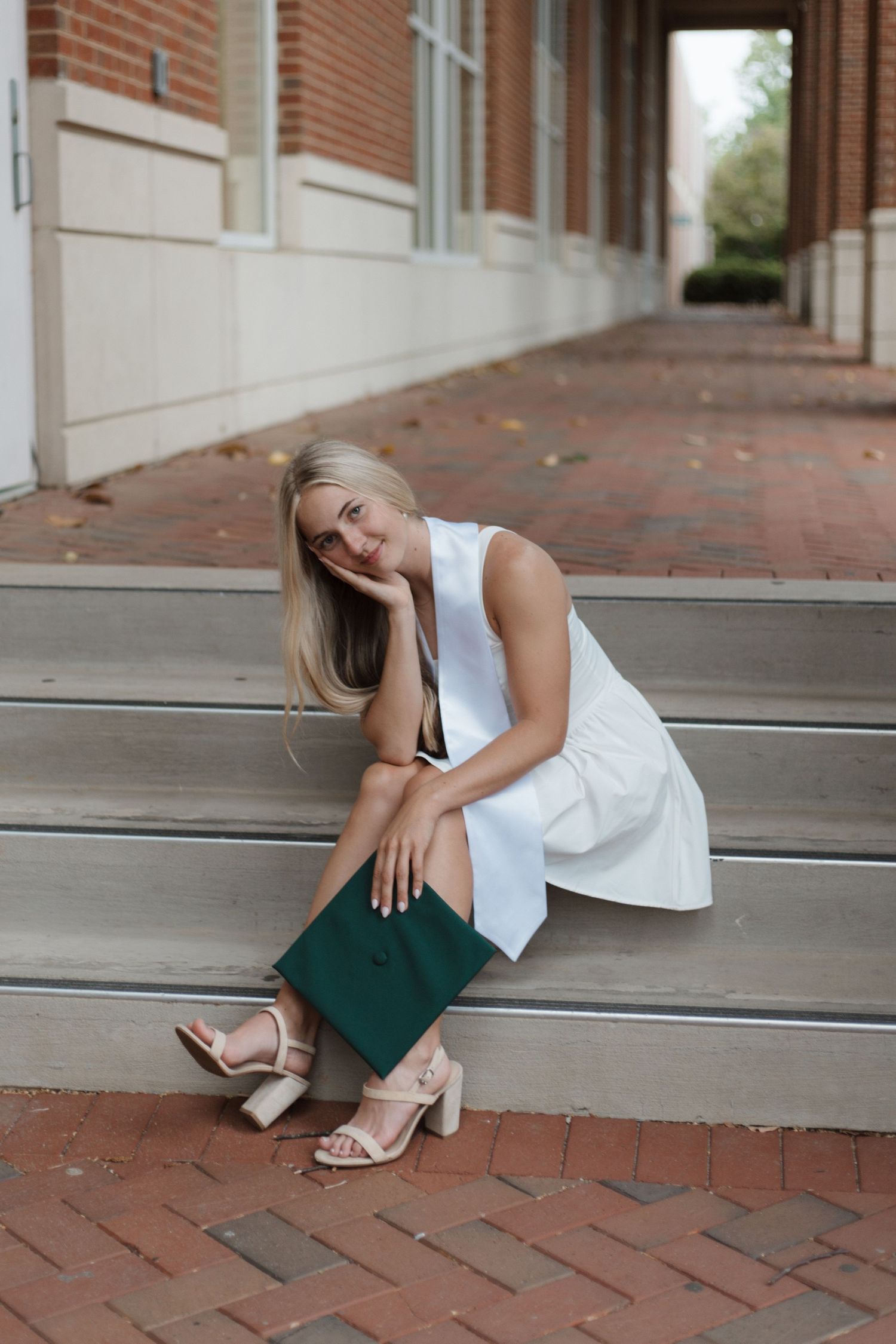 Young blonde woman in white dress holding green graduation cap, sitting on brick steps outside university building.