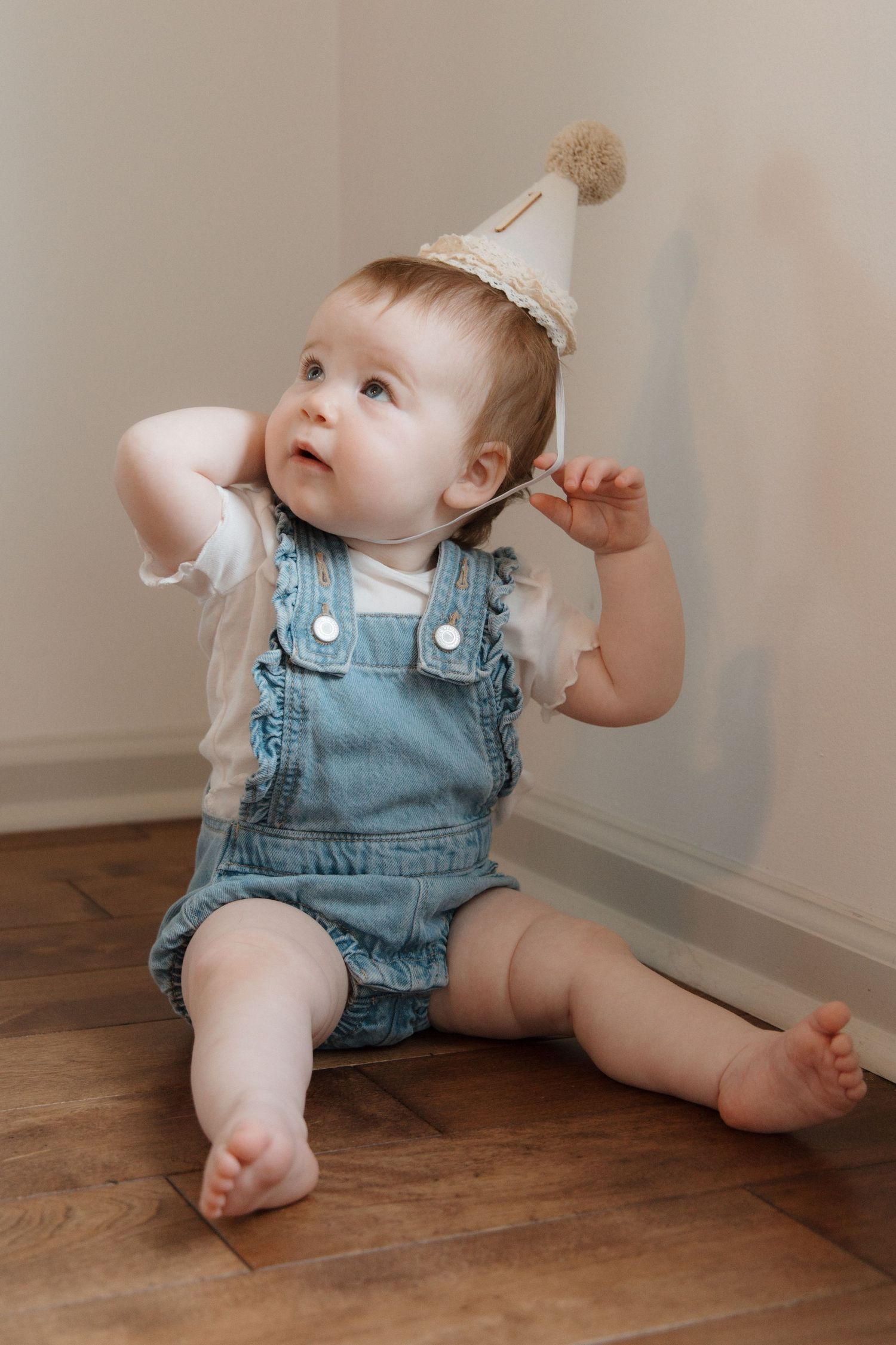 Adorable baby girl in denim overalls and birthday party hat sitting on hardwood floor looking up curiously.