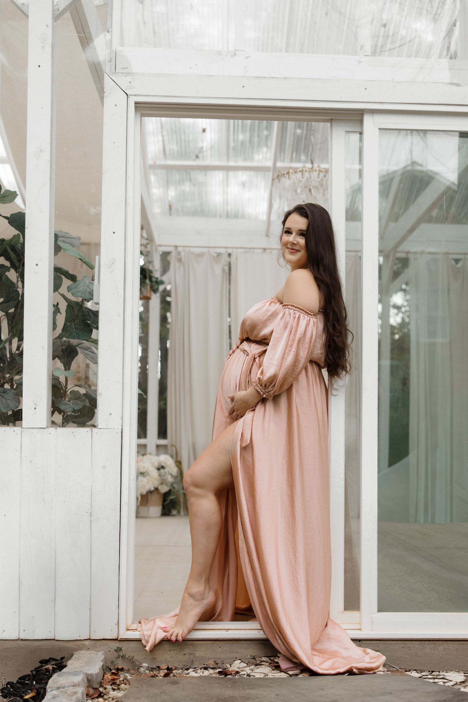 Pregnant woman in elegant blush pink off-shoulder maternity gown posing in a white greenhouse doorway.