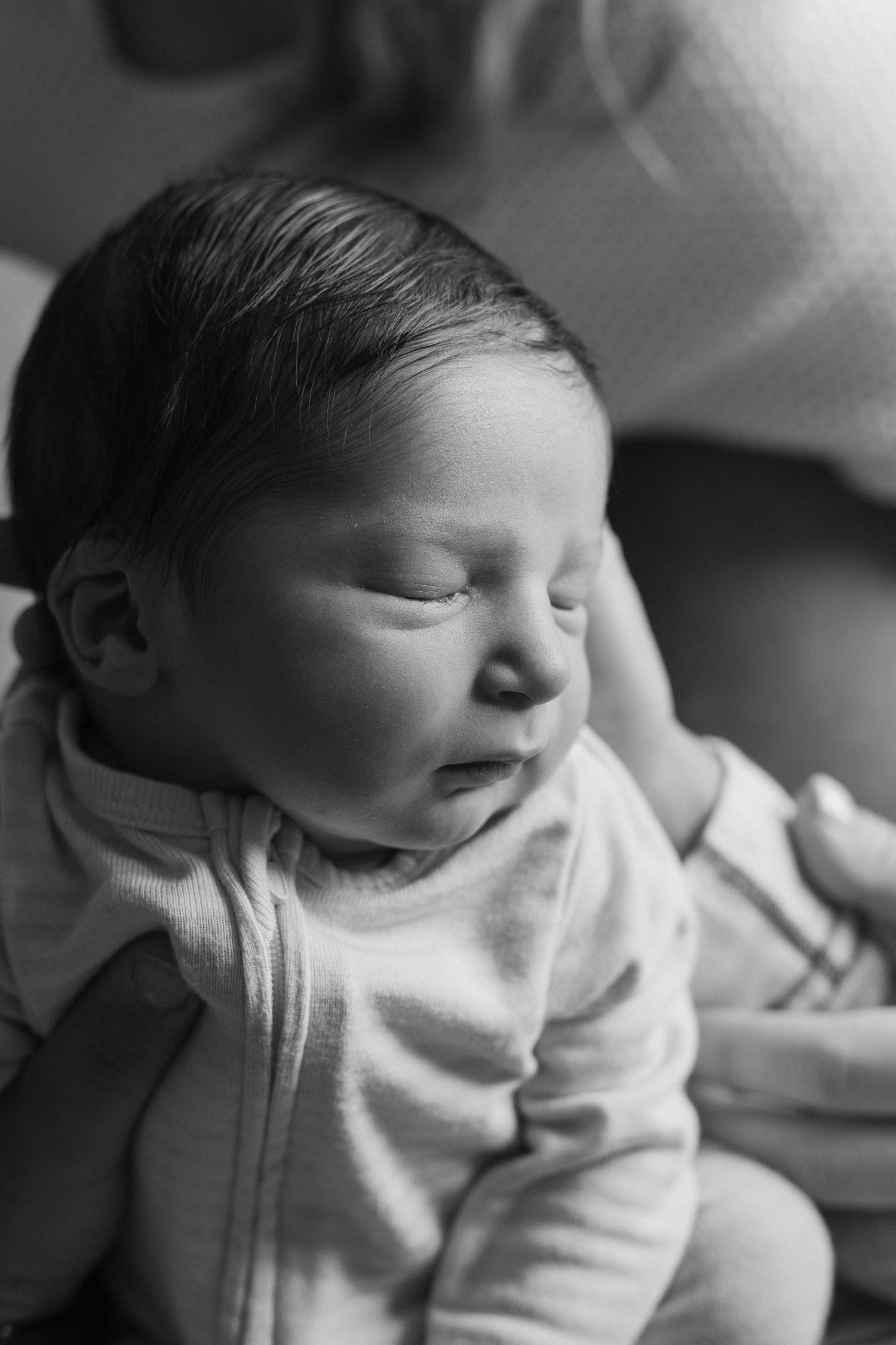 Black and white photo of a peaceful sleeping newborn baby with dark hair, dressed in a soft onesie.
