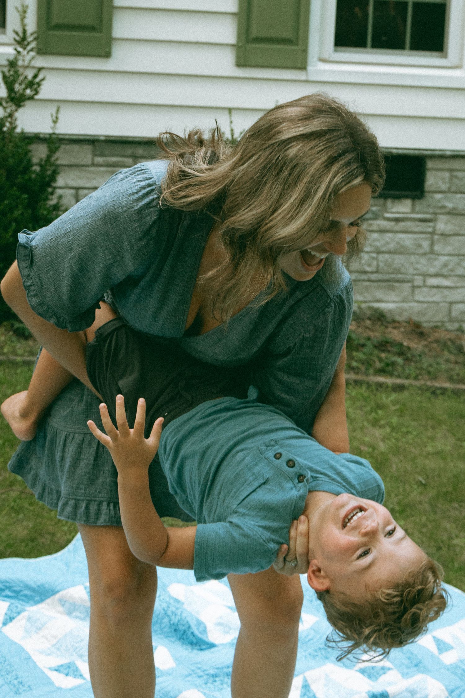 Woman in blue dress playfully holding laughing child upside down on blue quilt in backyard.