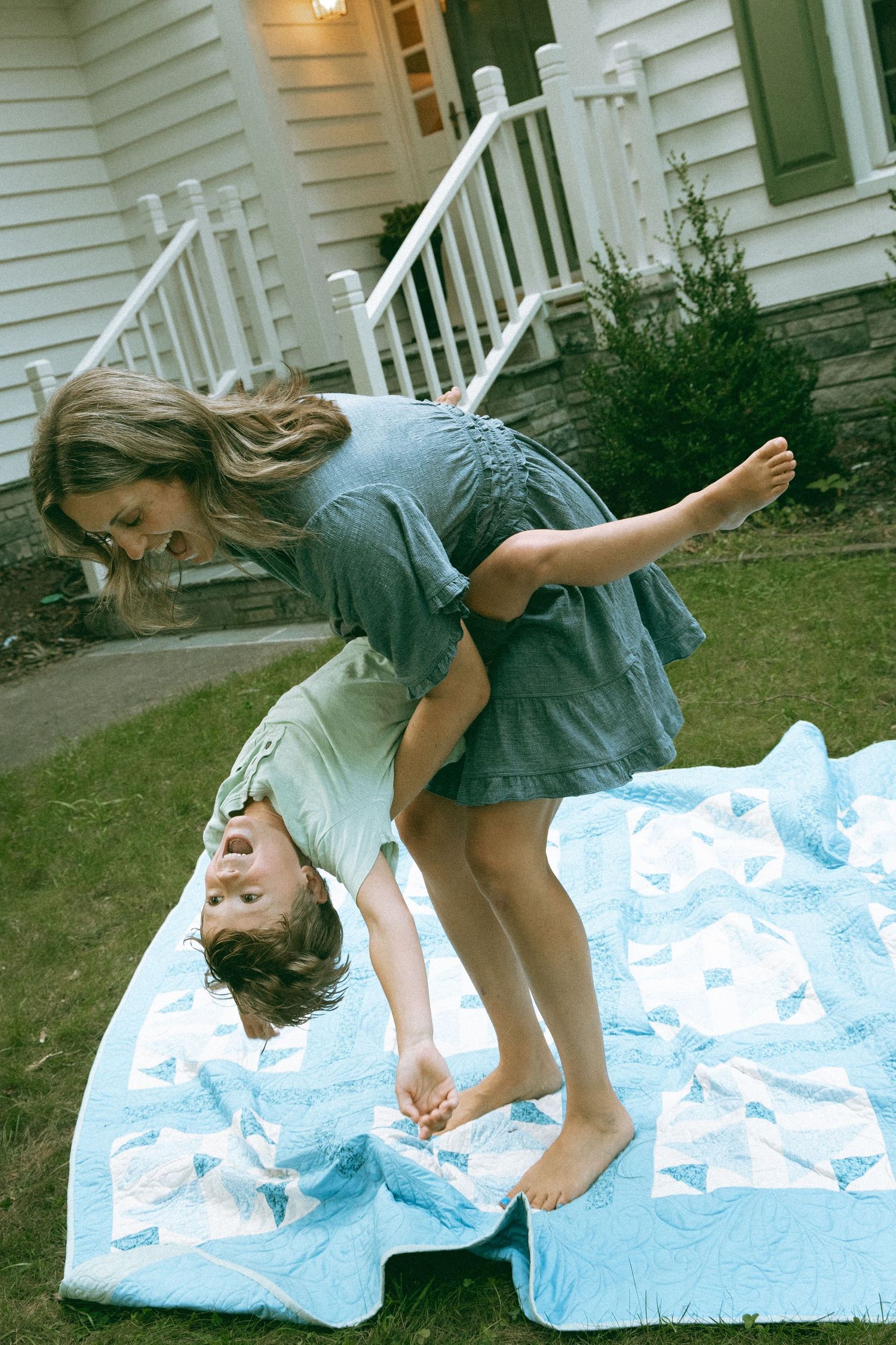 Mother in gray dress flipping giggling child upside down on blue patchwork quilt outside home.