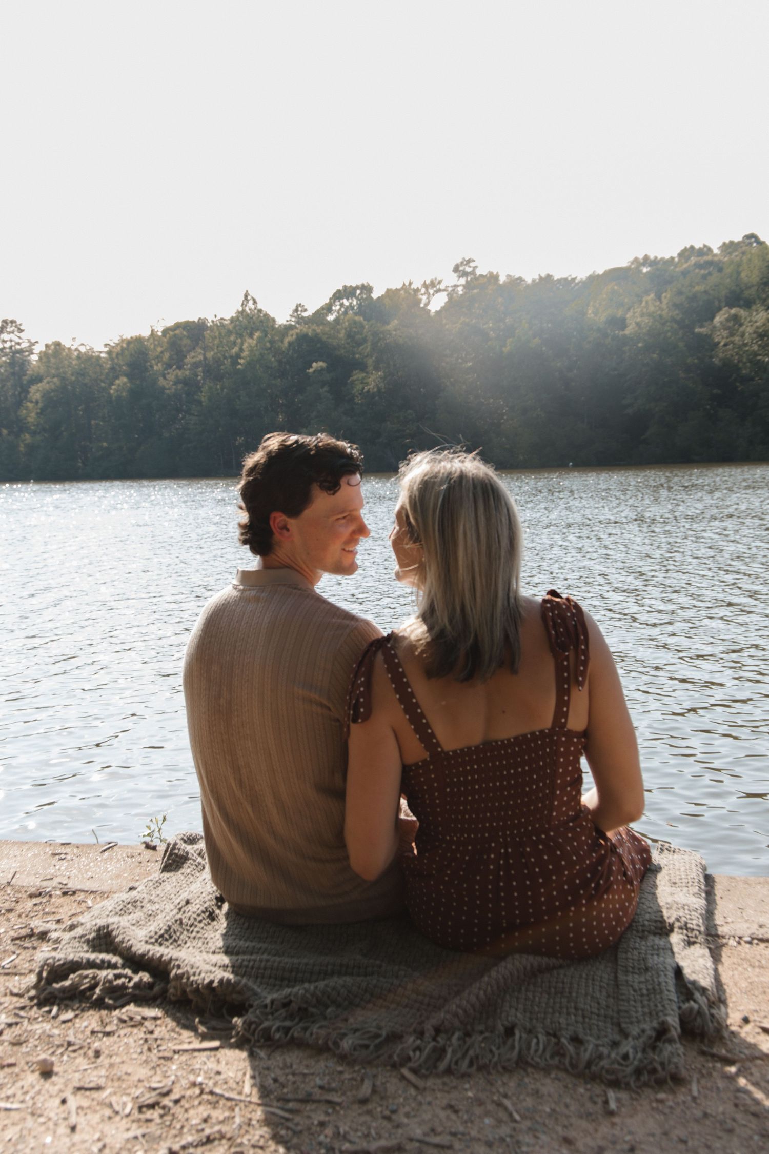 Couple sitting together on a blanket by a serene lake, surrounded by lush trees in warm golden sunlight.