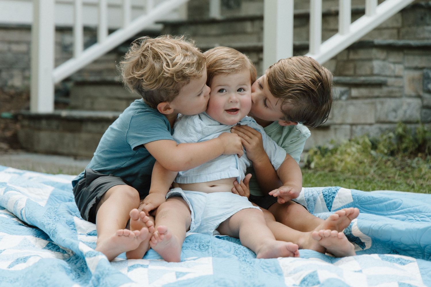 Three young children sitting on a blue quilt outdoors, two toddlers kissing a smiling baby on the cheeks.