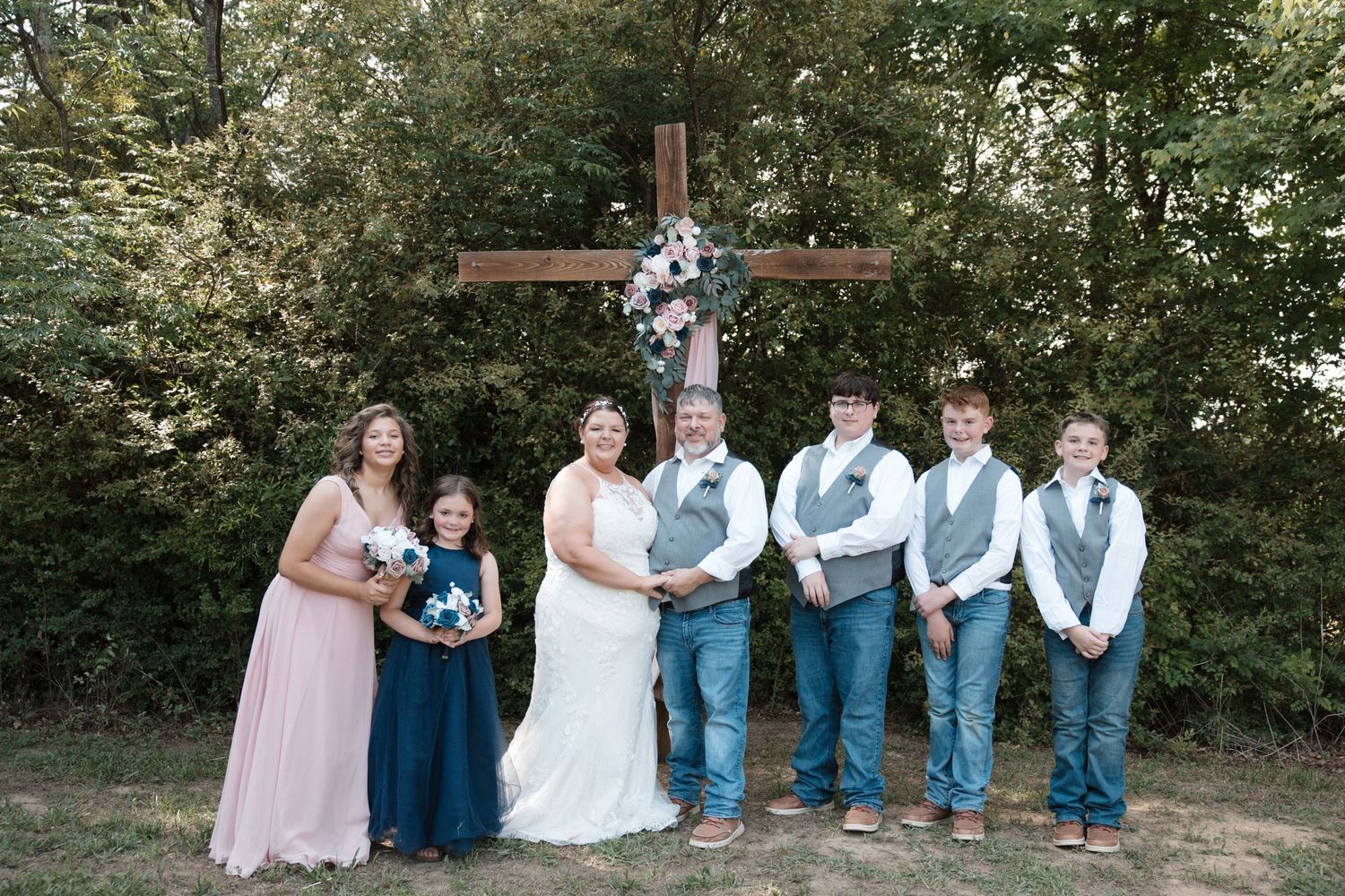 Wedding party poses before a wooden cross decorated with flowers, featuring bride, groom, groomsmen in vests, and bridesmaids outdoors.