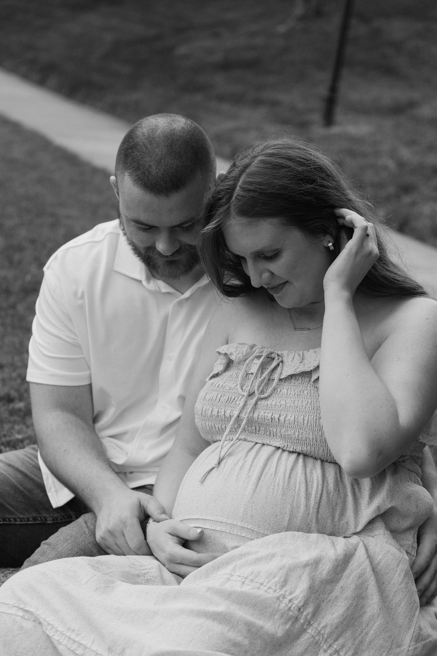 Black and white photo of a smiling couple sitting outdoors, lovingly looking down at the pregnant woman's baby bump.