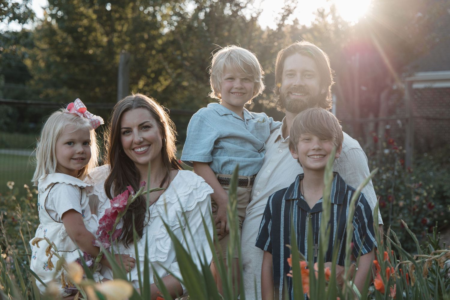 Family of five smiling outdoors at golden hour, with lush greenery and warm sunlight in the background.