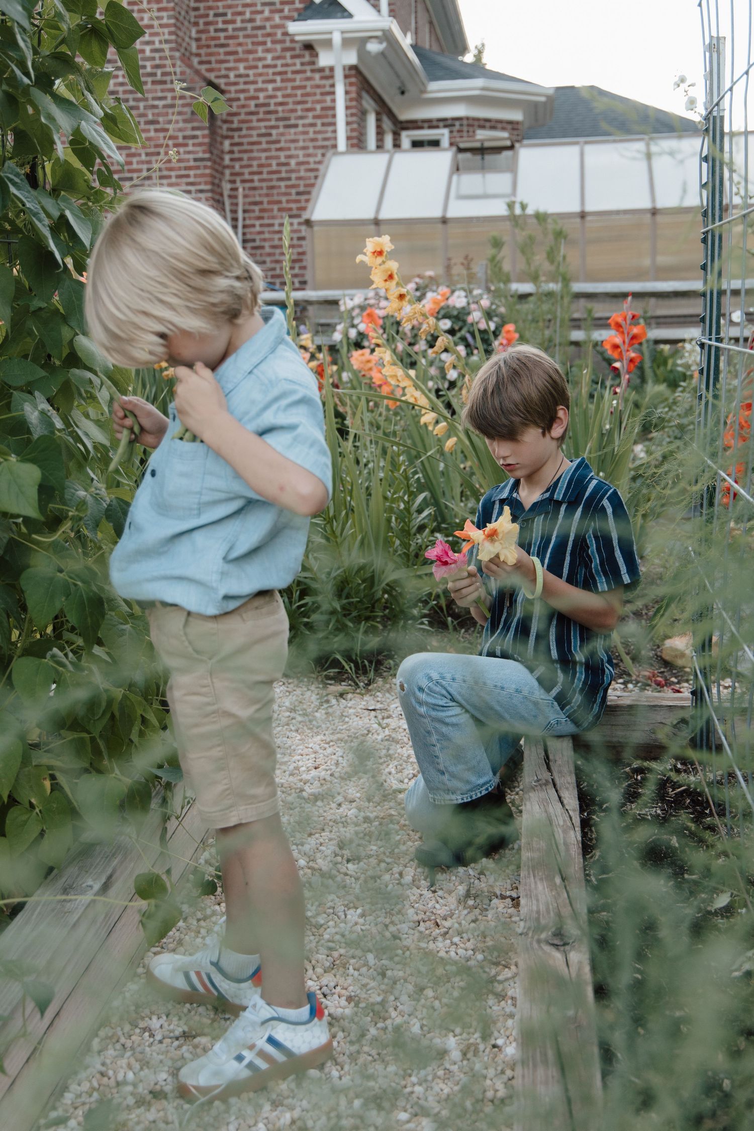 Two boys exploring a garden, examining flowers and plants near wooden raised beds with a brick building in the background.