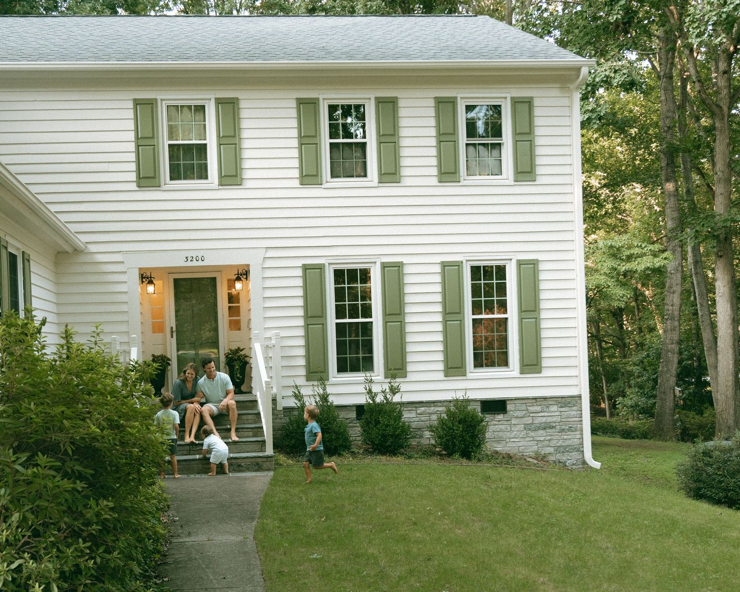 White two-story colonial home with green shutters, manicured lawn, and family gathered on front porch steps.