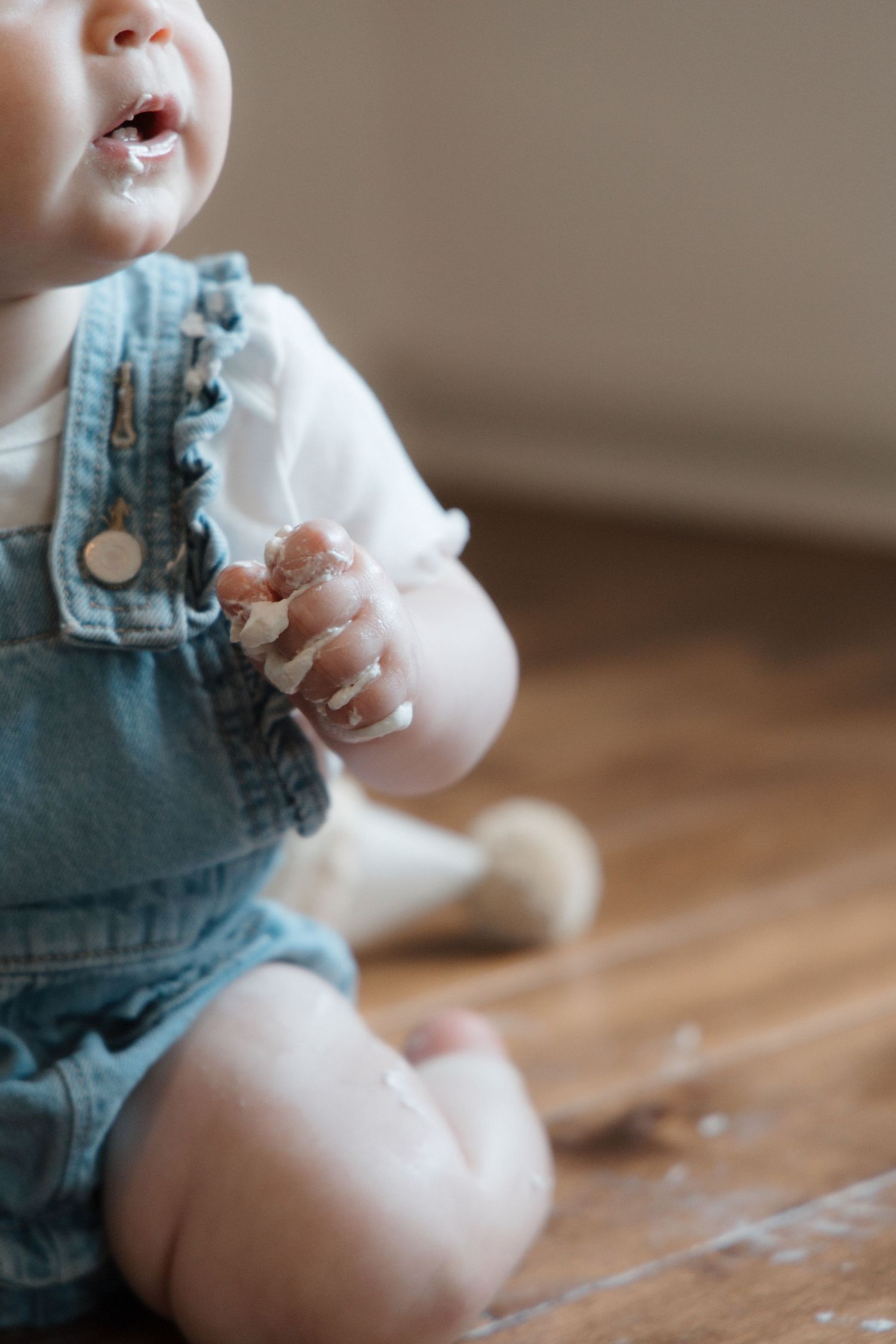 Baby in denim overalls sitting on hardwood floor with frosting on hands and mouth.