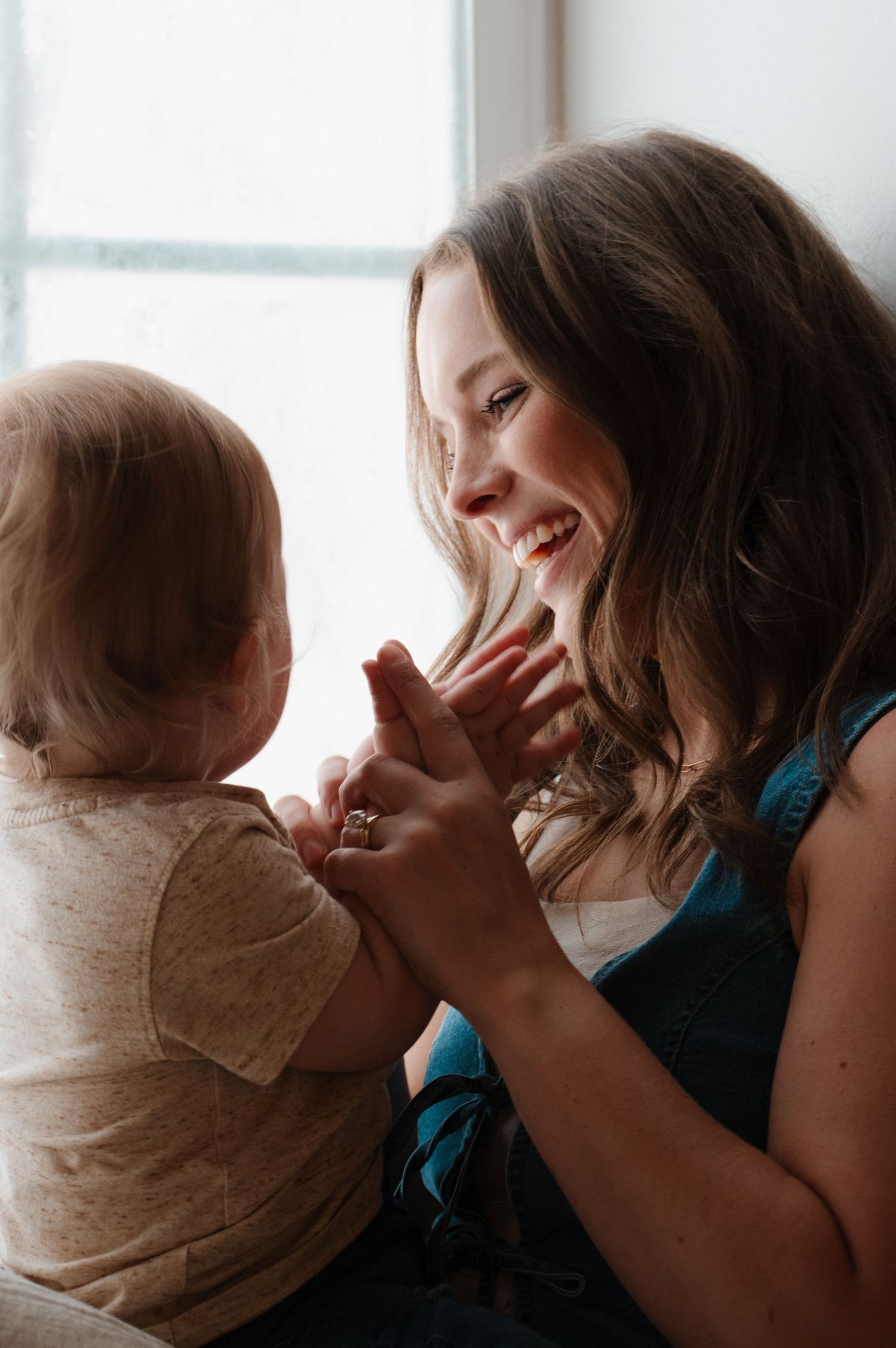 A smiling mother joyfully plays with her baby near a bright window, holding hands in a warm, loving moment. CHARLOTTE NC